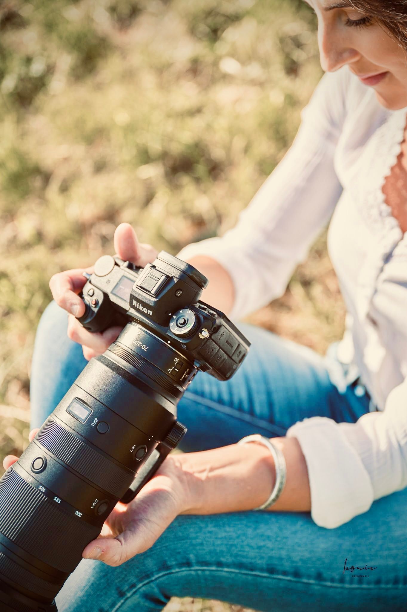Woman in white shirt holding a large Nikon camera with telephoto lens outdoors in natural light.