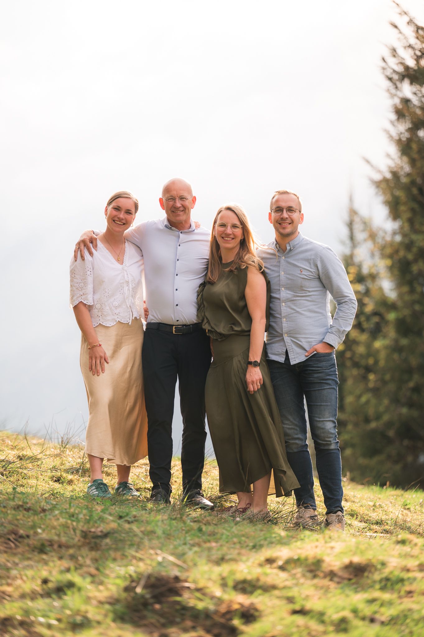 Family of four smiling outdoors on a grassy hillside, posing together in casual attire on a cloudy day.