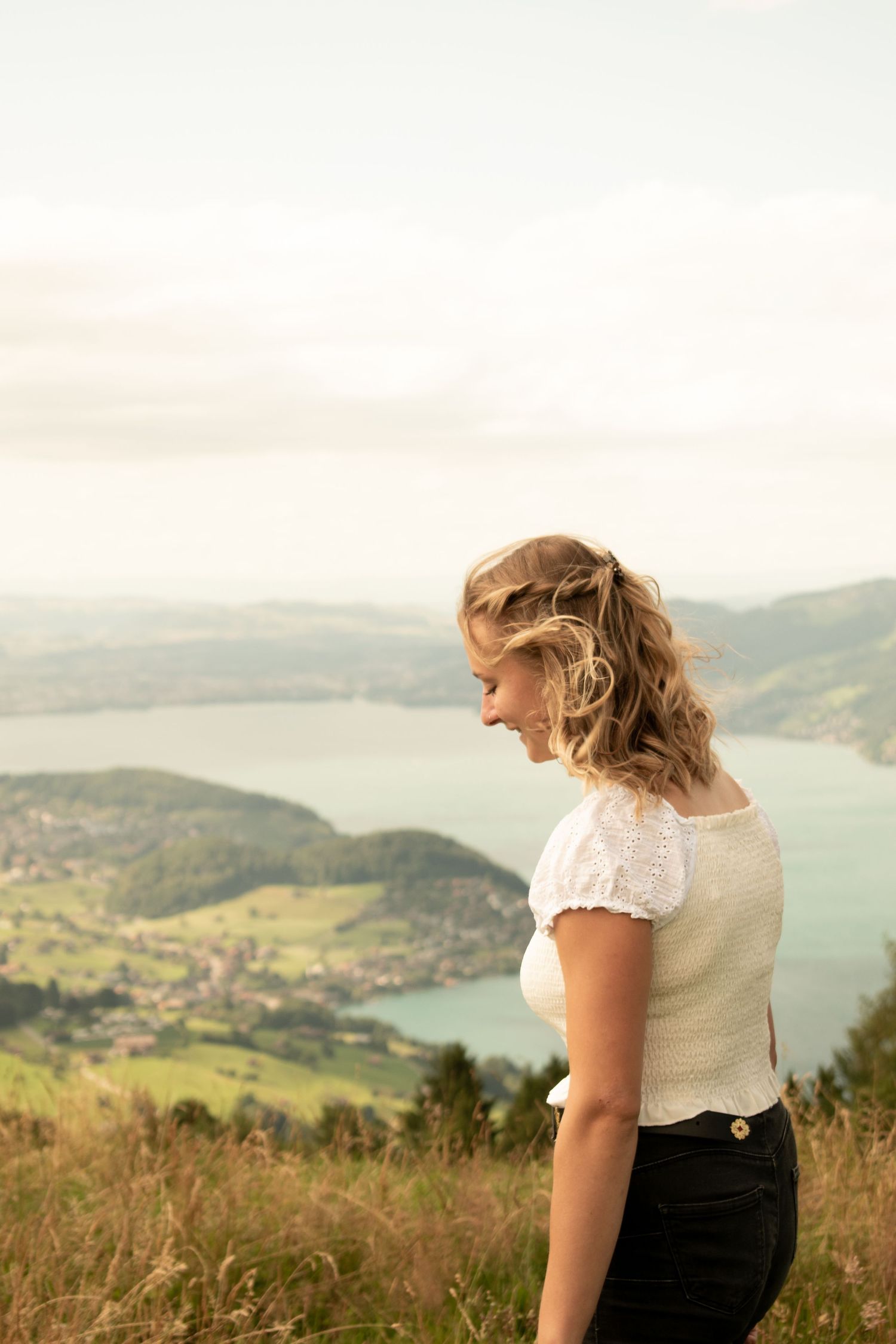 Person in white lace top looking out over scenic mountain lake view from hilltop.