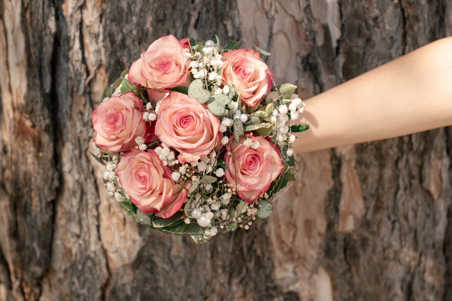 A hand holds a delicate bouquet of pink roses and baby's breath flowers against a rustic tree bark background.