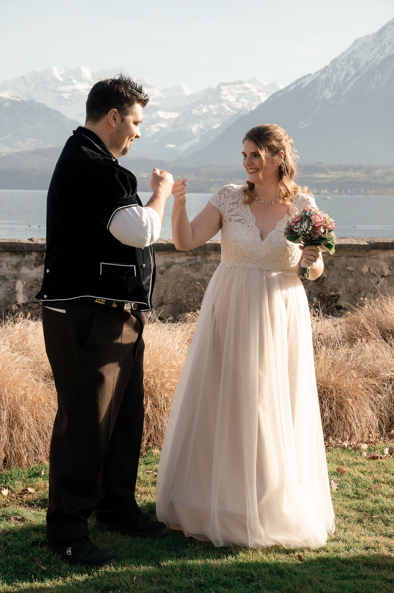 Couple in wedding attire dances together on a hilltop with snow-capped mountains in the background at sunset.