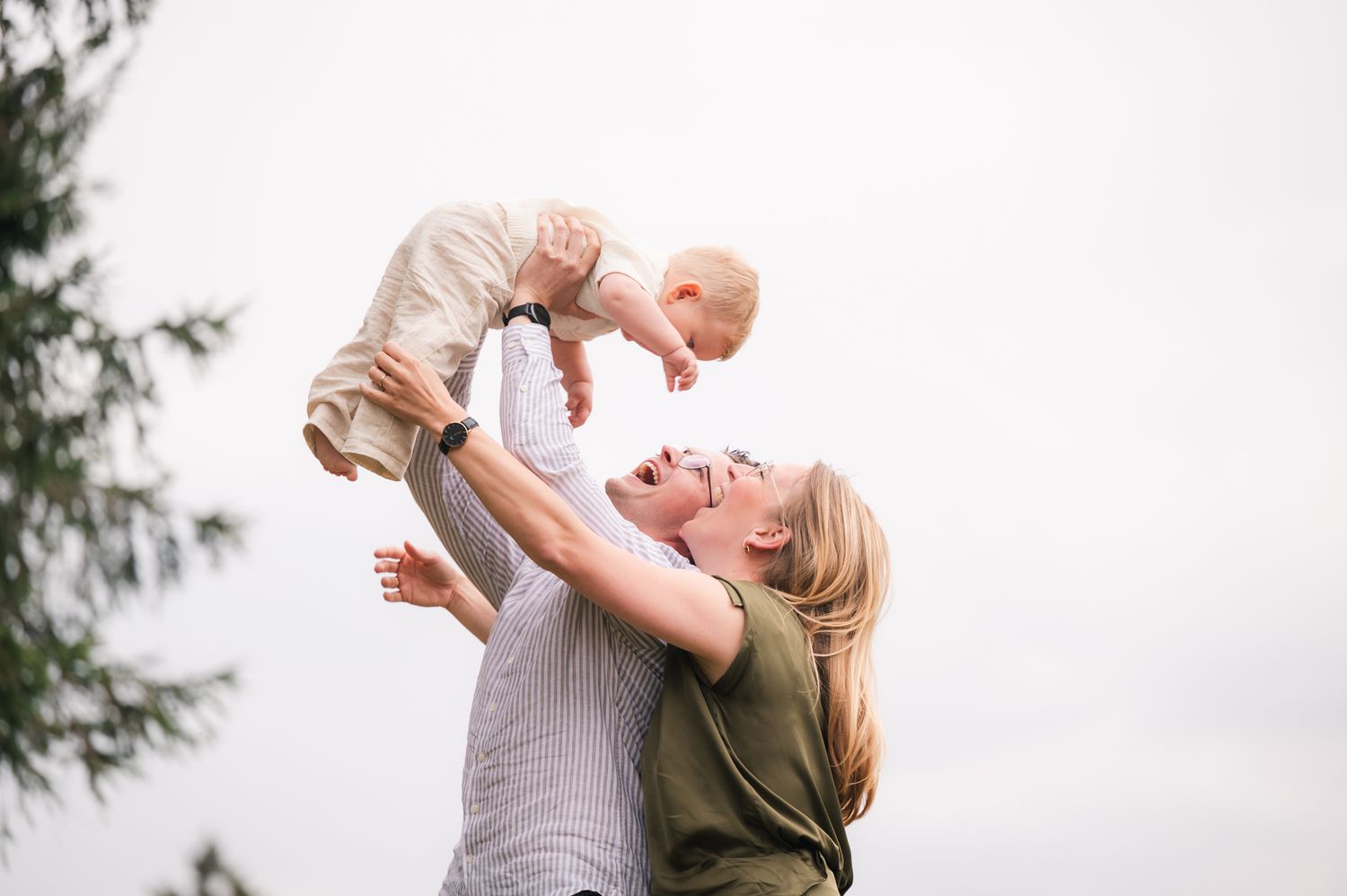 A joyful mother lifts her smiling baby into the air outdoors, both dressed casually in a heartwarming moment.