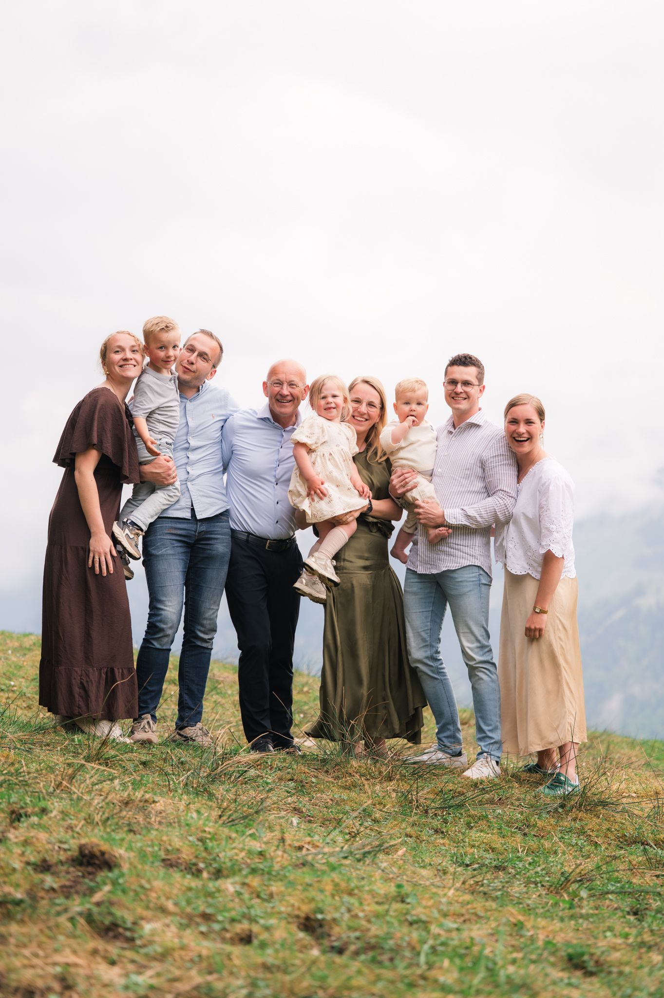 Multi-generational family portrait outdoors on a grassy hillside, smiling together on a cloudy day.