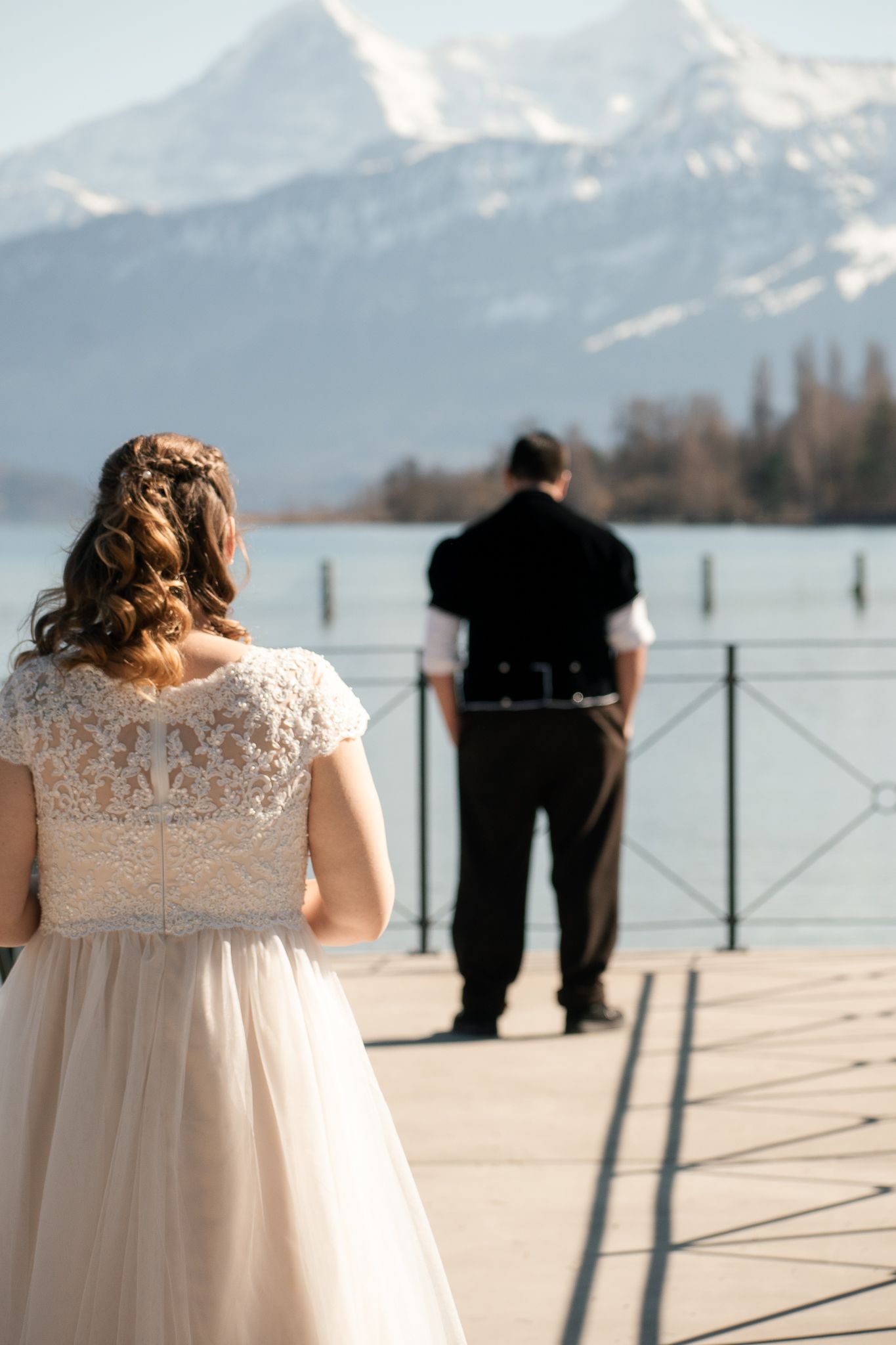 People gather on a lakeside deck with snowy mountains in the background on a sunny day.