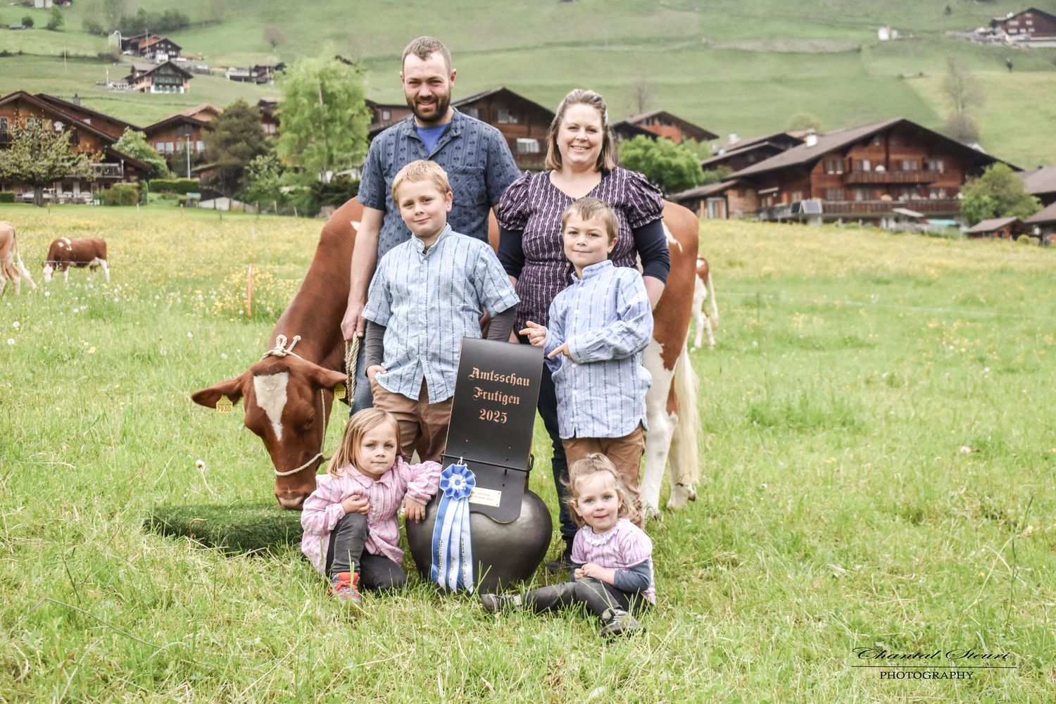 Group gathered around milk can in scenic Alpine meadow with traditional wooden chalets.