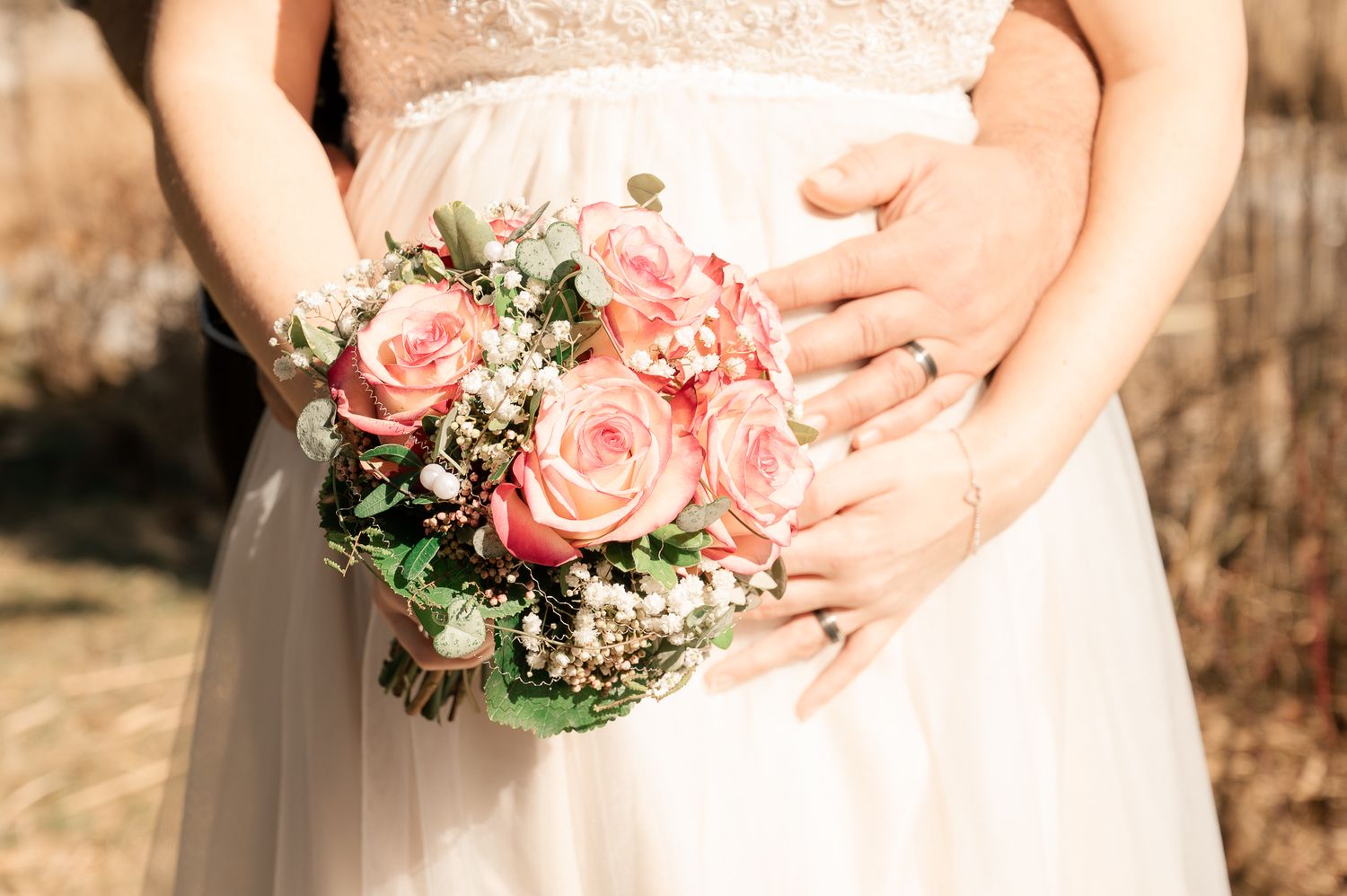 A wedding couple holds a pink and white floral bouquet against a rustic outdoor backdrop.