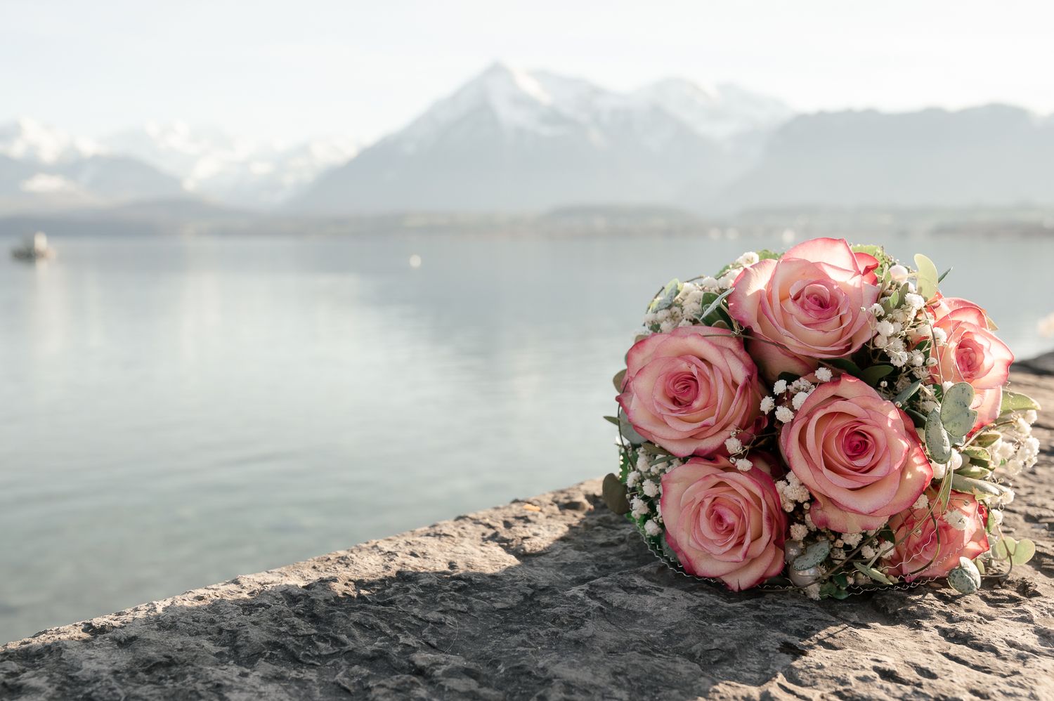 Pink roses and floral bouquet rests on rocky shoreline with misty mountain lake view in background.
