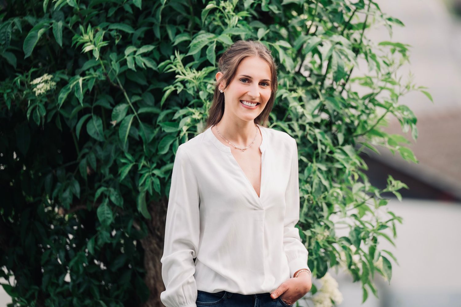 Smiling woman in white blouse standing outdoors against lush green foliage backdrop.