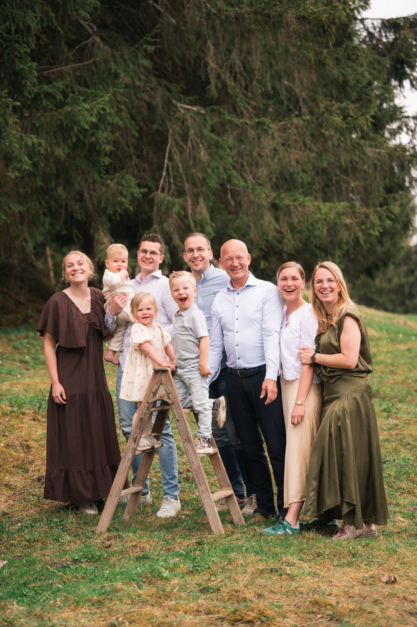 Large family group portrait outdoors on grass, smiling together near a wooden ladder with evergreen trees behind.
