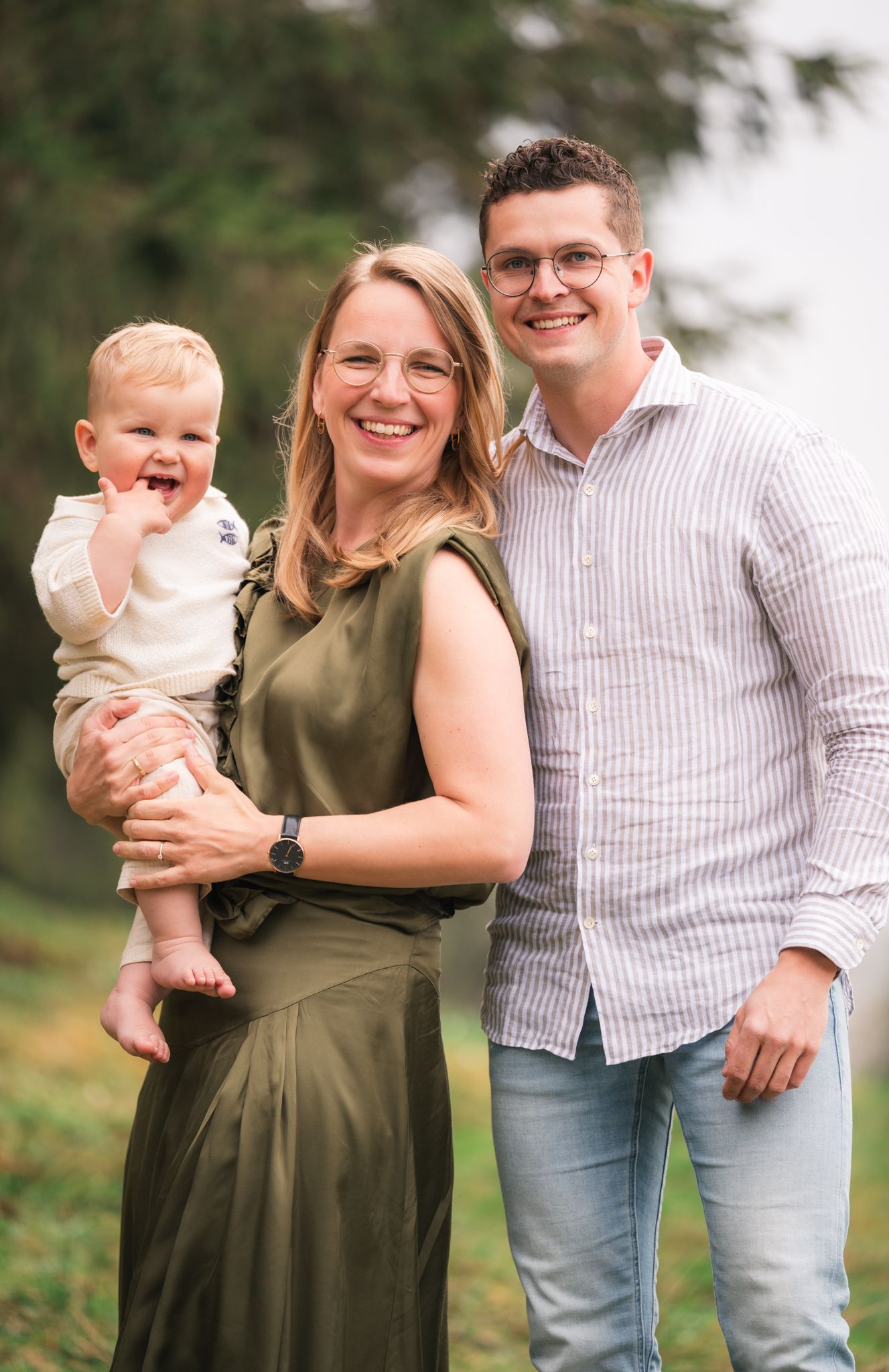 A smiling family of three poses outdoors, with a woman in a green dress holding a happy baby beside a man in a striped shirt.