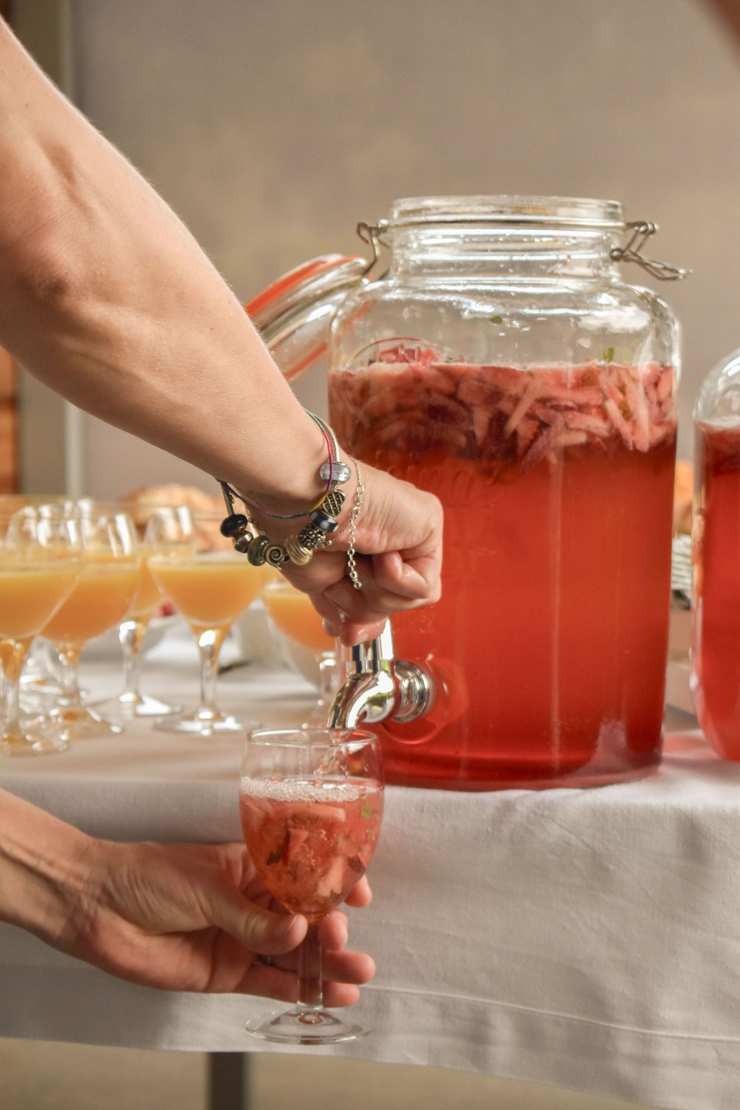 Hands pouring pink cocktails from a large glass dispenser at an event.