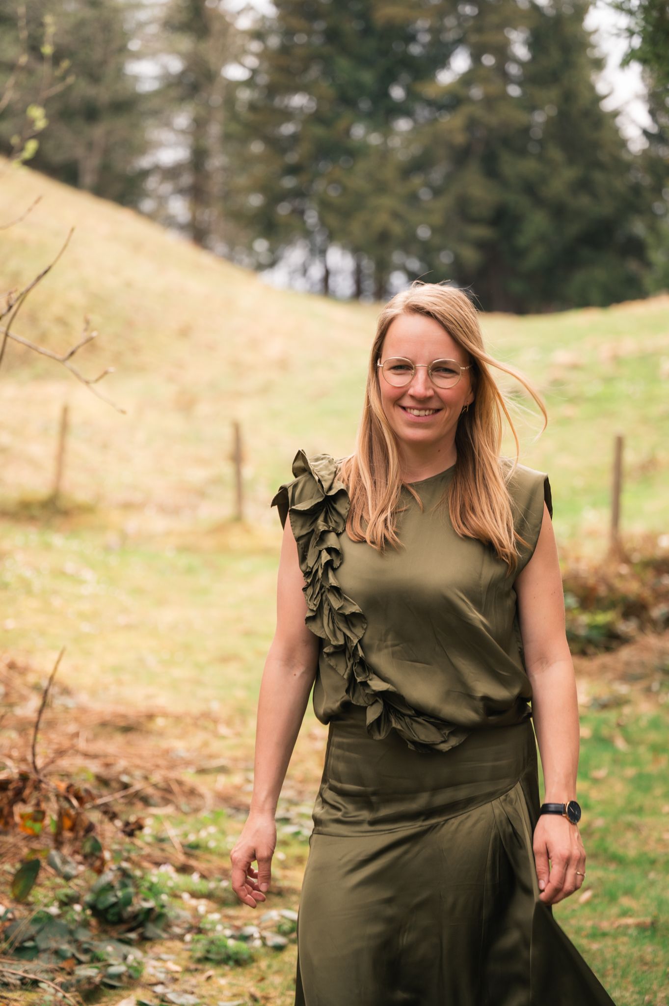Smiling blonde woman in olive green ruffled dress standing outdoors on a green hillside with trees in background.