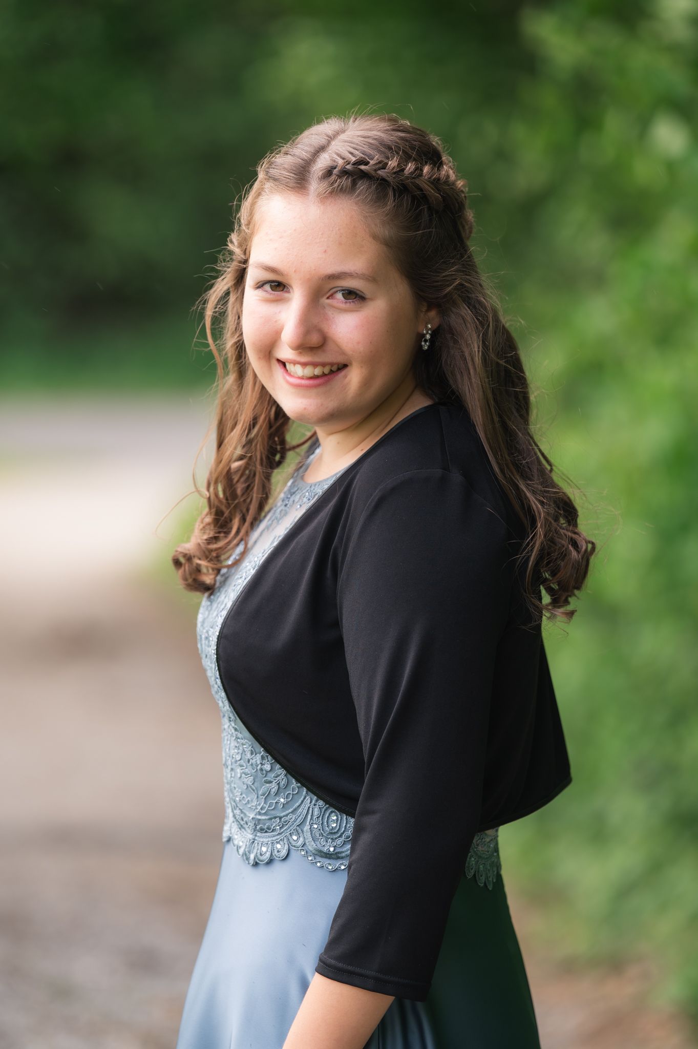 Young woman in blue gown and black bolero jacket smiling outdoors with braided hairstyle.