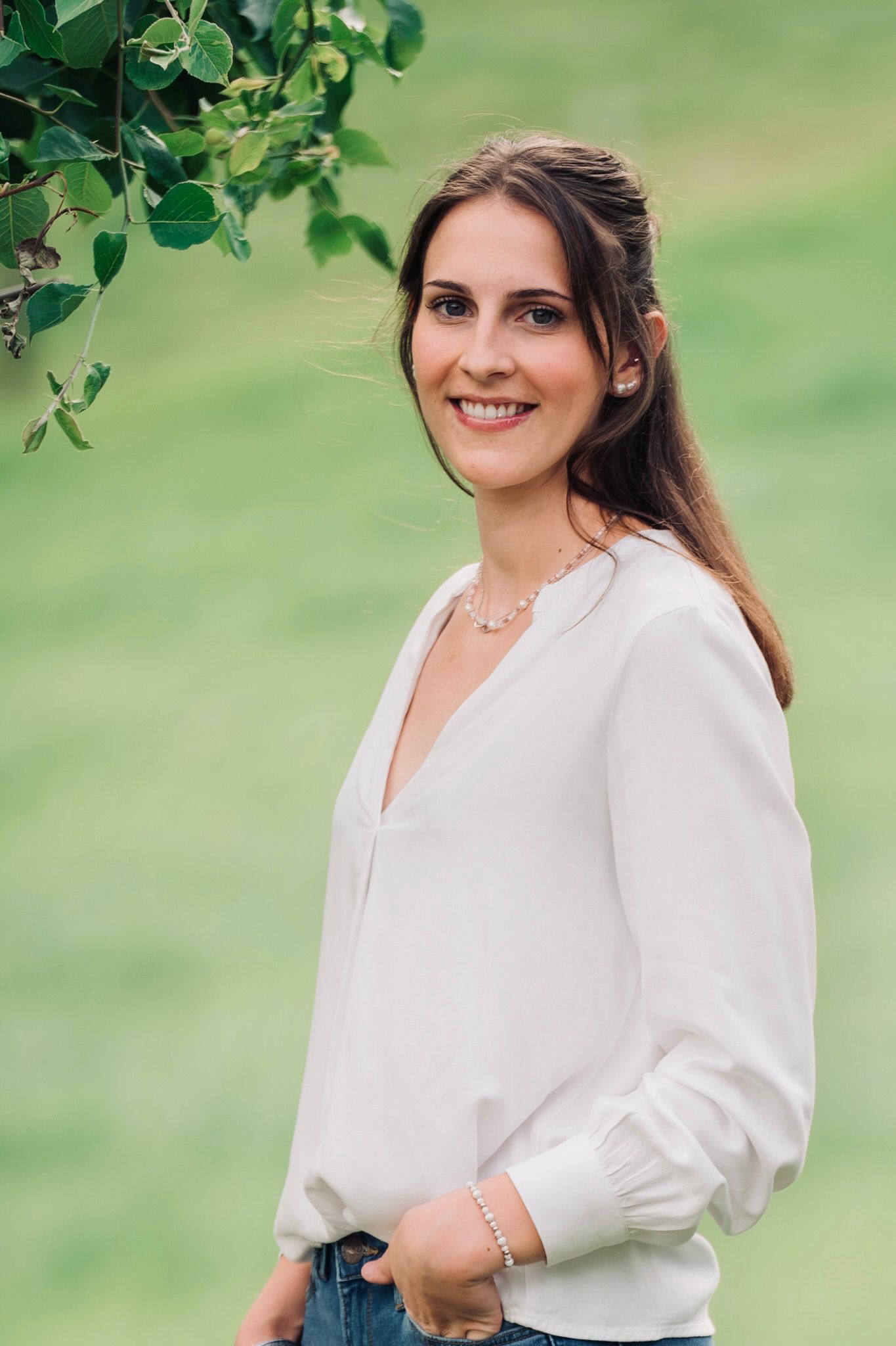 Smiling young woman in white blouse and jeans standing outdoors near green tree branches on a sunny day.
