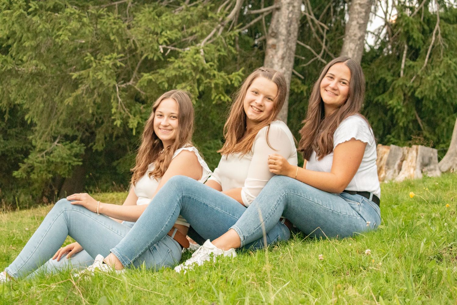 Three friends lounging on green grass wearing coordinated white tops and blue jeans.