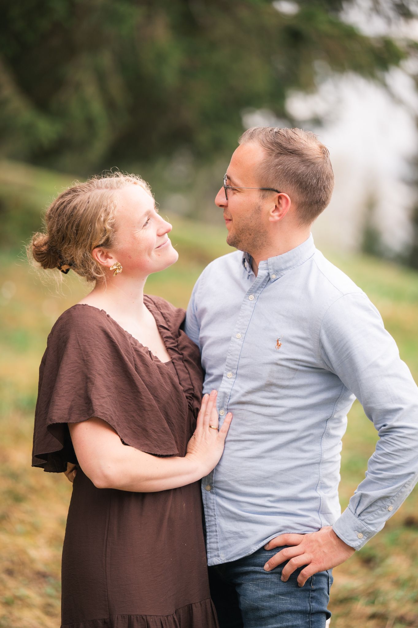 A couple sharing a romantic moment outdoors, woman in brown dress gazing up at man in light blue shirt.