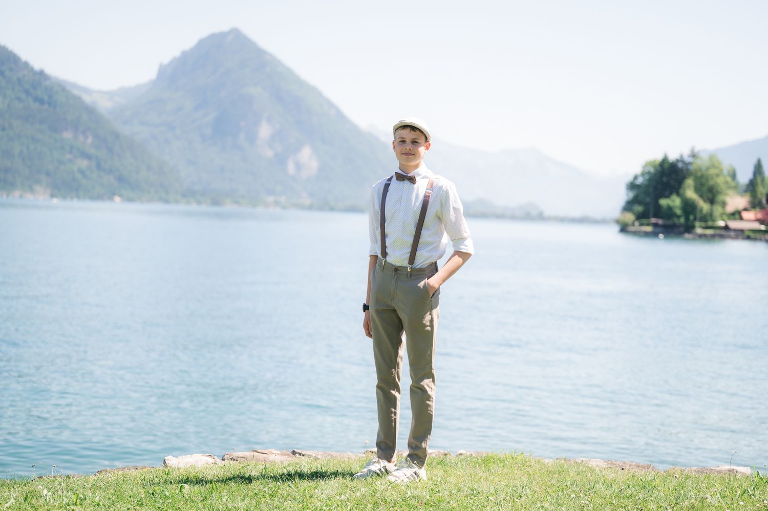 Man in suspenders and bow tie stands by a serene mountain lake on a sunny day.