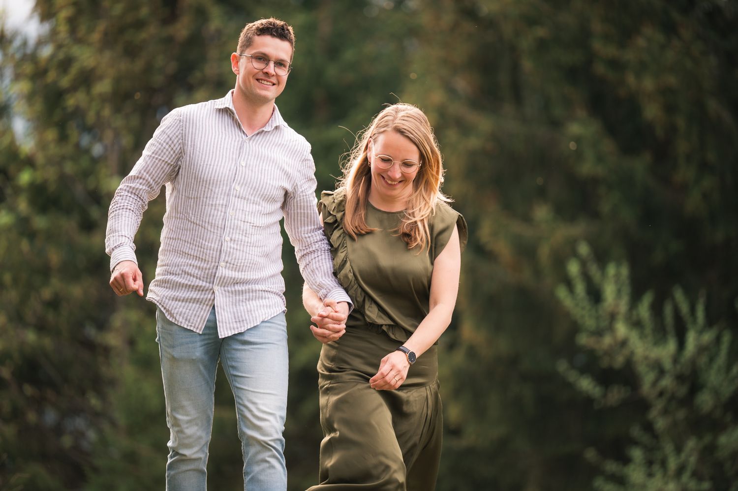 Couple holding hands and walking through a lush green park, smiling and enjoying a sunny outdoor engagement photo session.