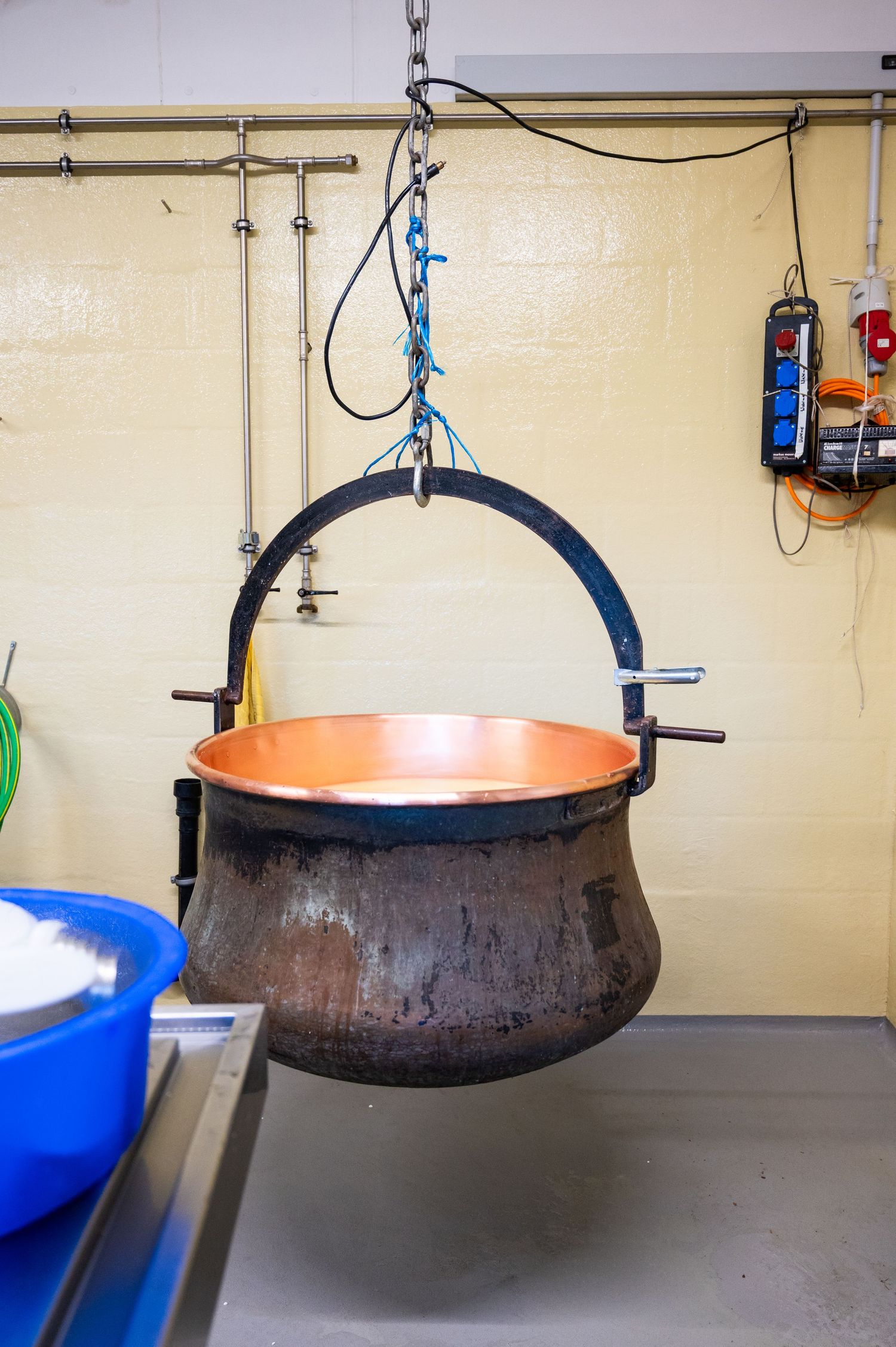 Large copper cauldron hanging from chains in a workshop or industrial kitchen setting.