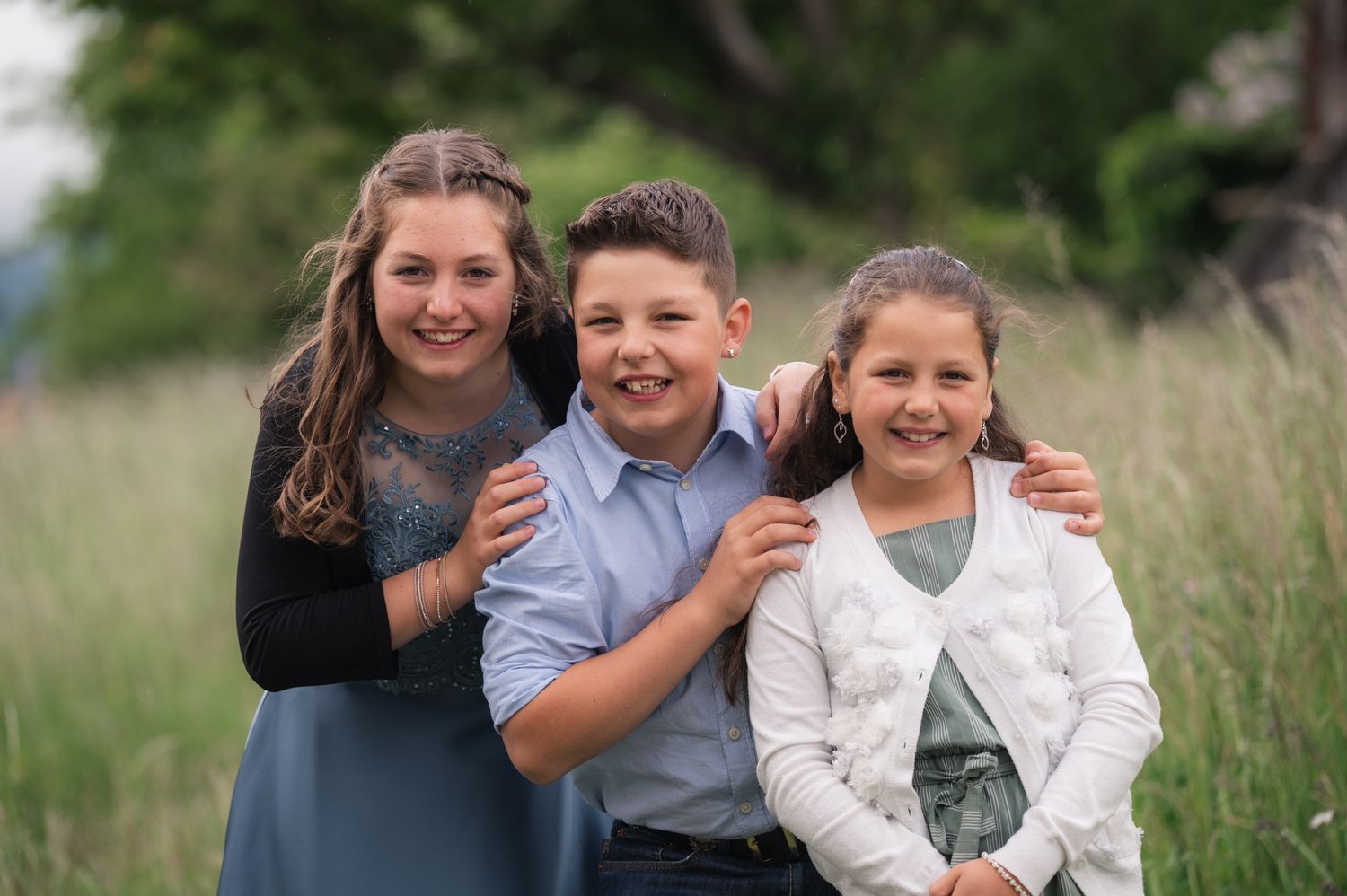 Three smiling children pose together in a grassy field, wearing semi-formal attire for an outdoor family portrait.