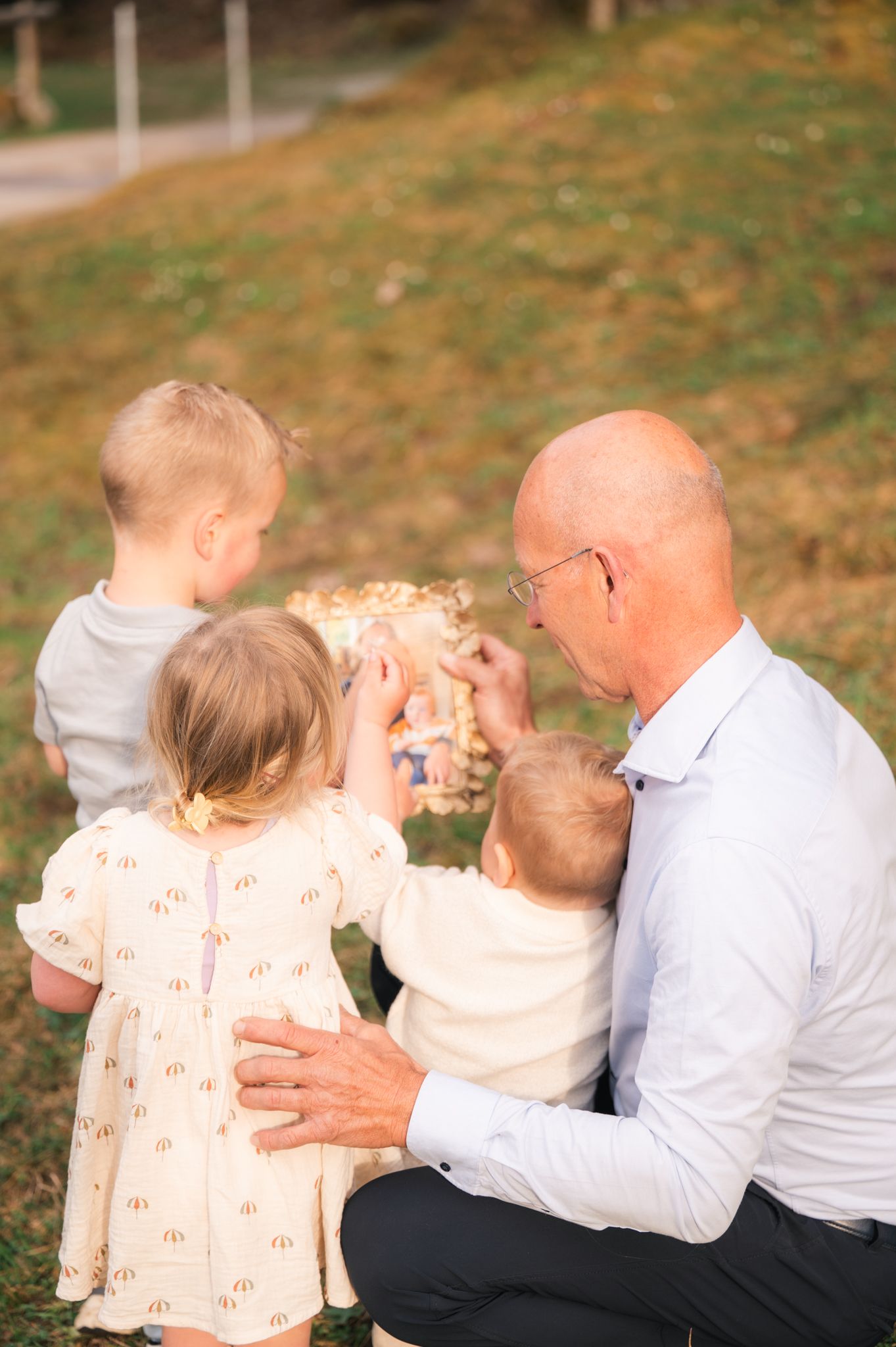 Grandfather crouching outdoors with three young children, sharing a joyful moment while holding a toy in a green park.