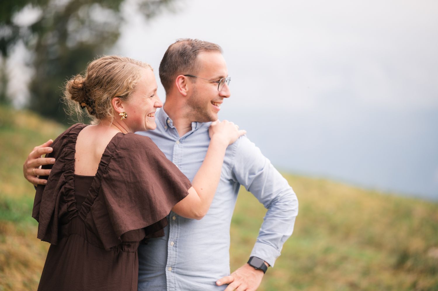 Couple embracing outdoors on a grassy hillside, woman in brown wrap and man in light blue shirt smiling together.