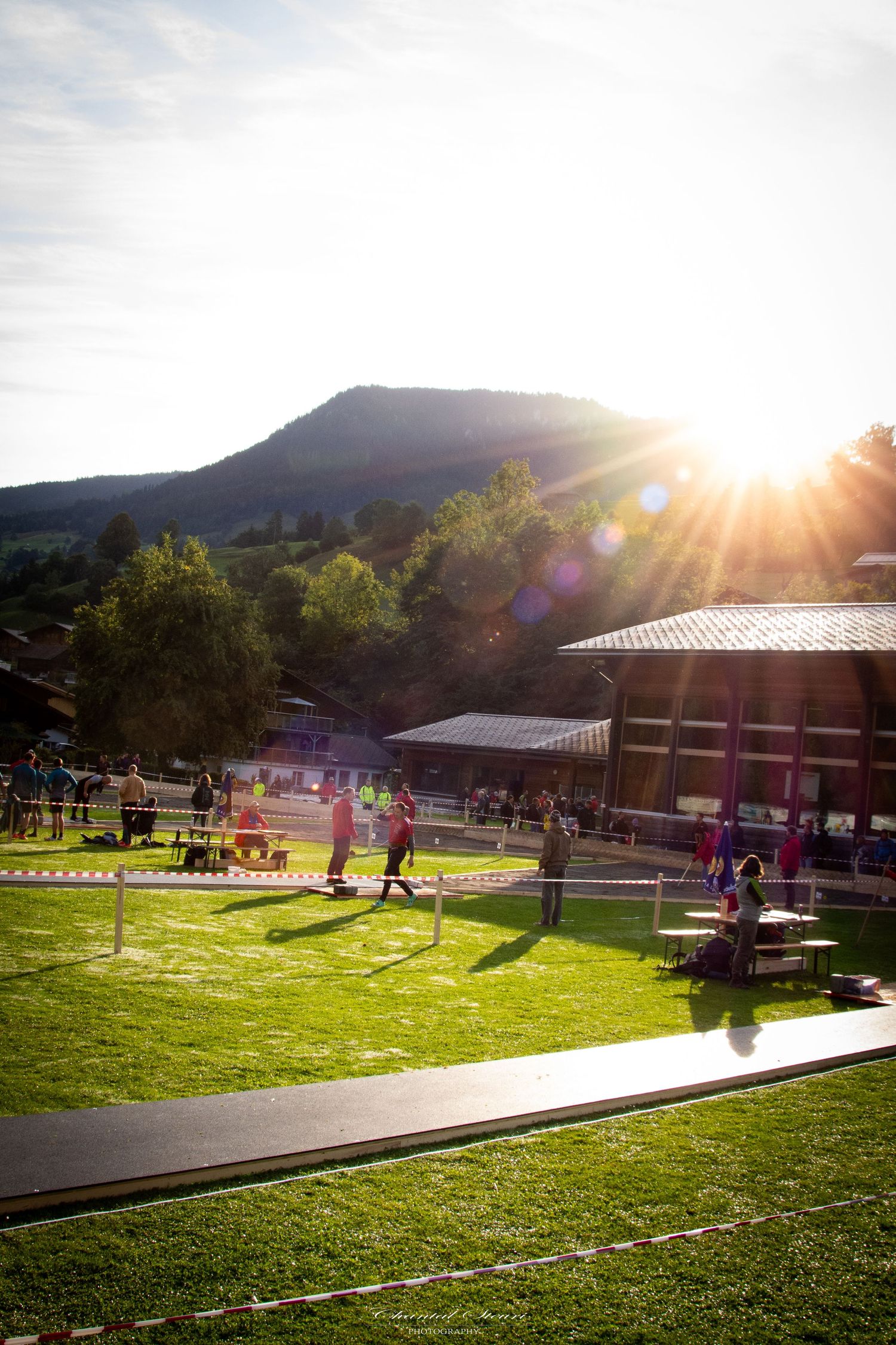 Sunset over mountain landscape with people enjoying outdoor activities on green lawn.