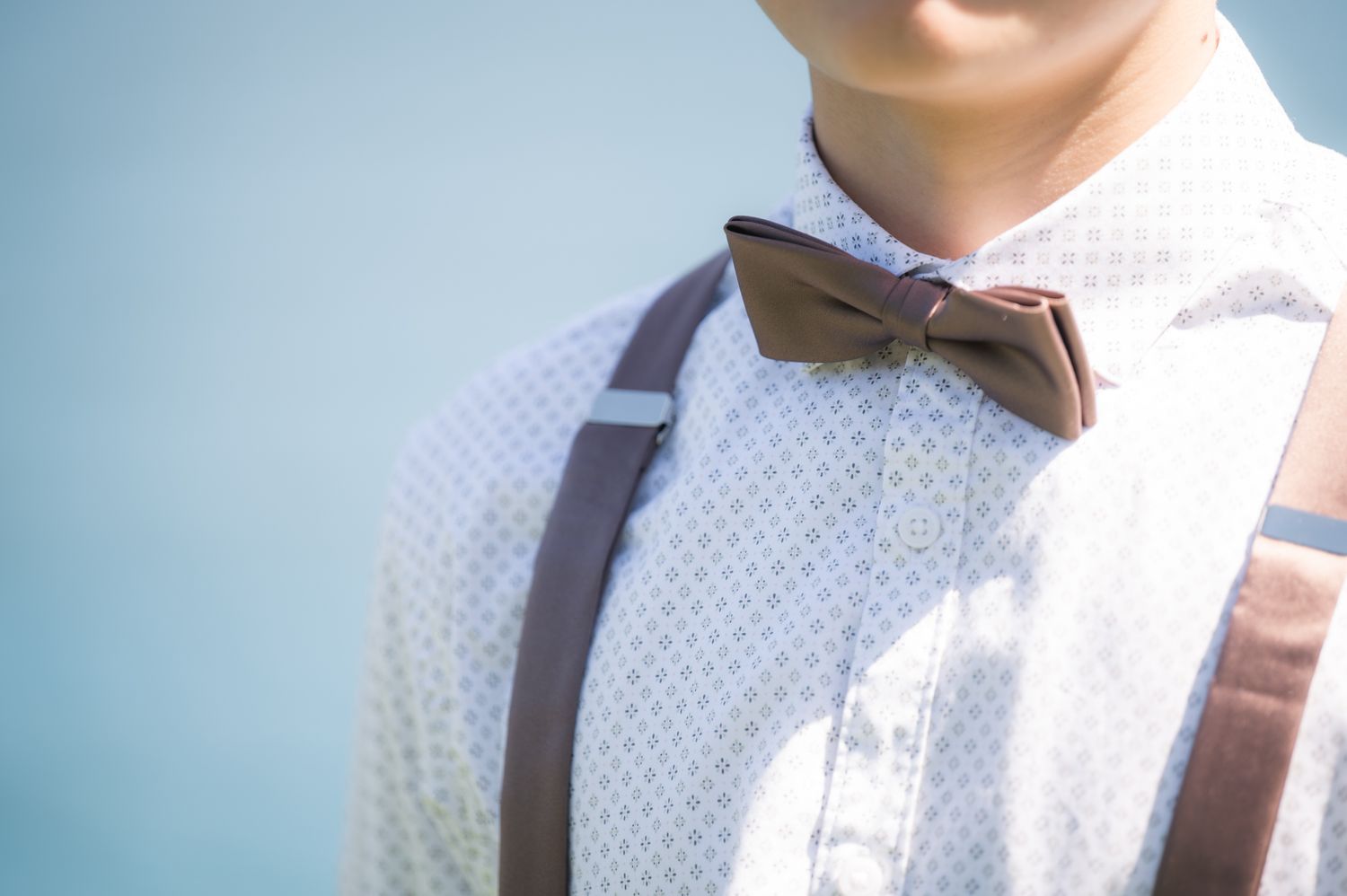 Young man wearing a brown bow tie, suspenders, and a white patterned dress shirt against a light blue background.
