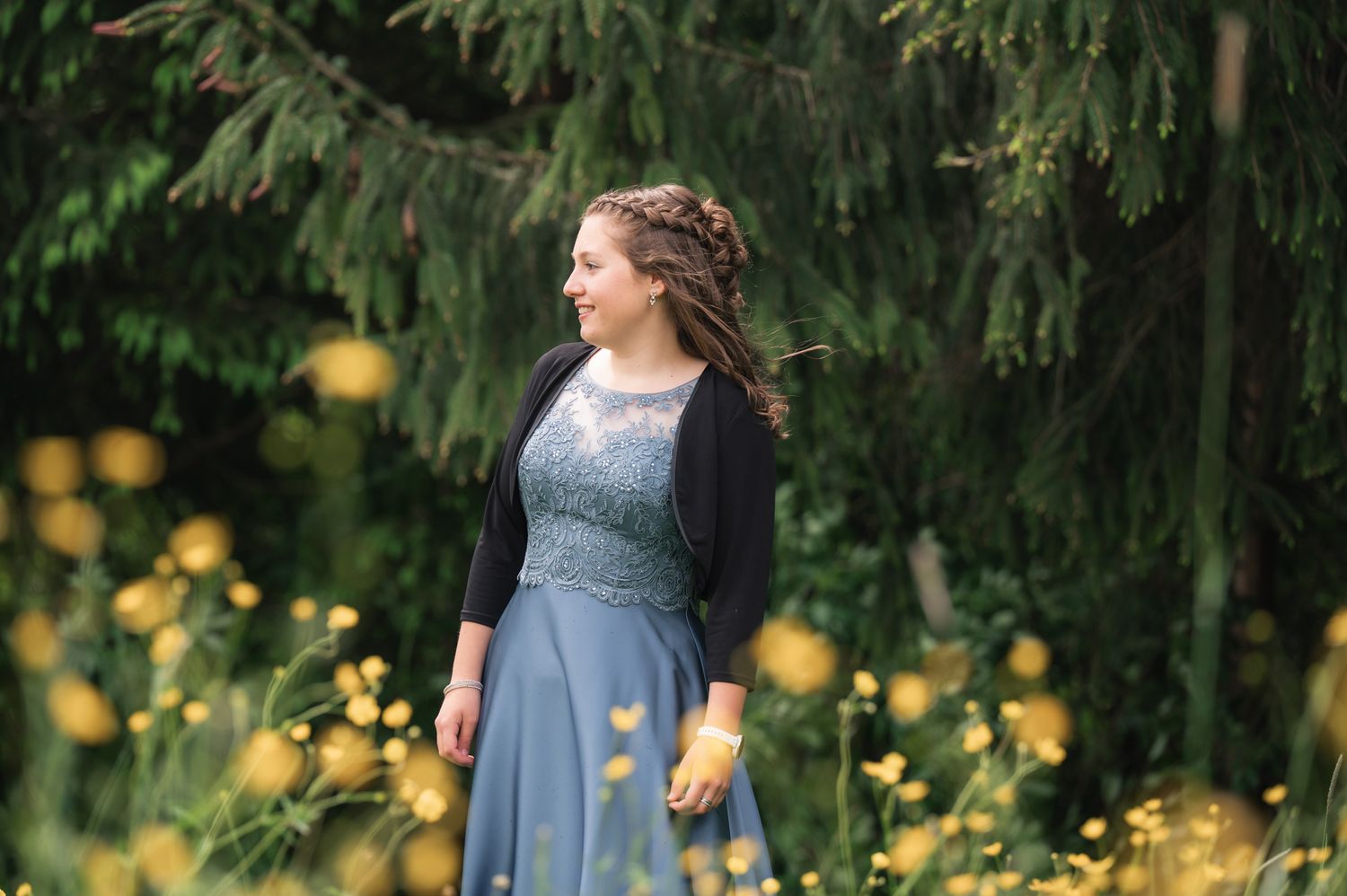 Young woman in blue gown and black shrug stands among yellow wildflowers with evergreen trees backdrop.