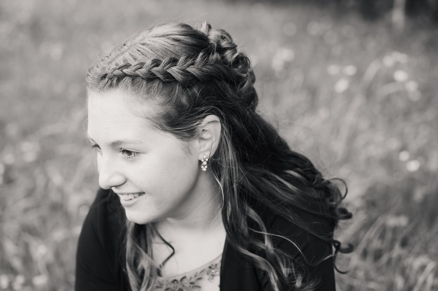 Black and white portrait of a young girl with braided half-up hairstyle, smiling outdoors in a field.