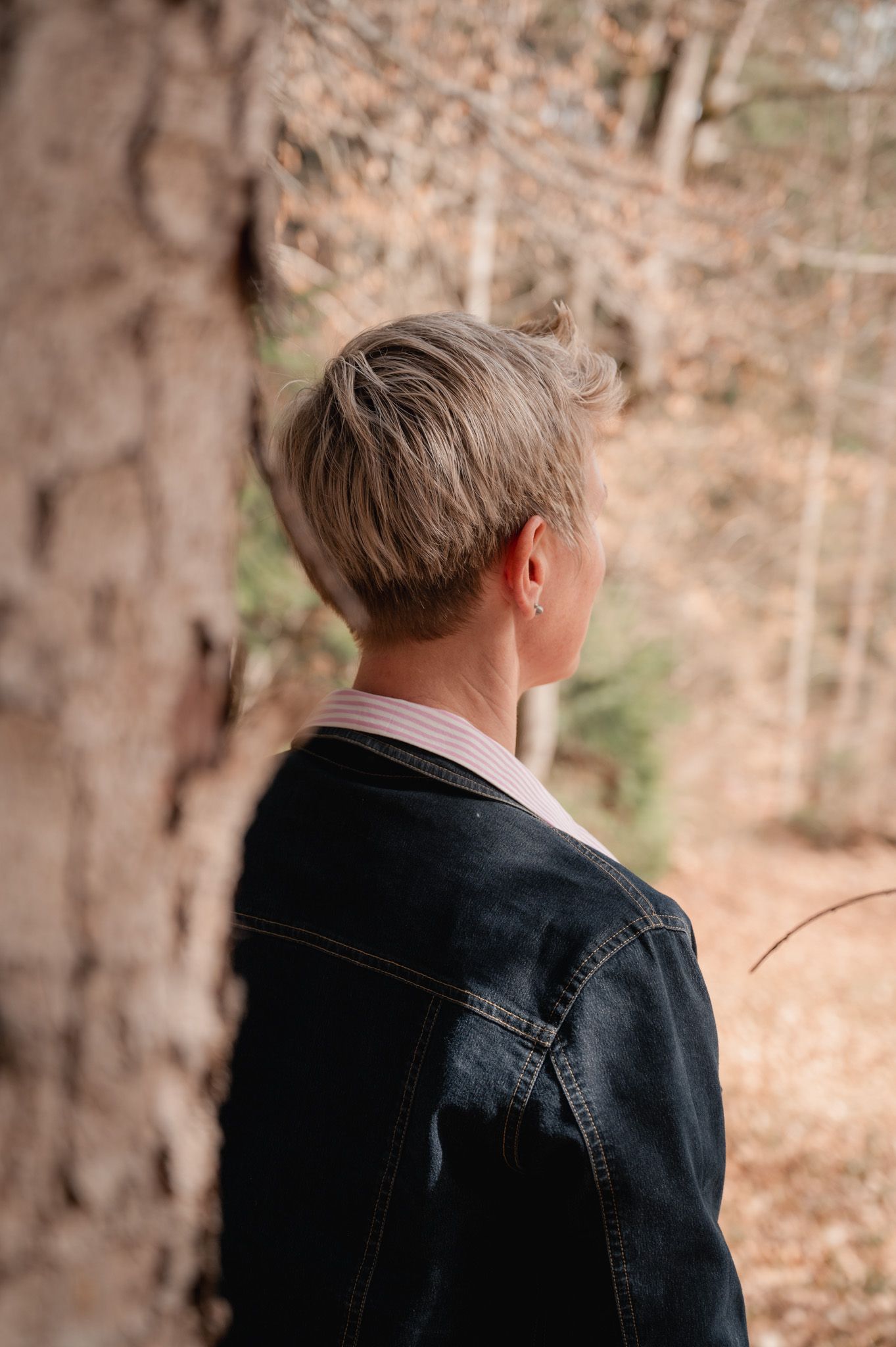 Back view of person with short blonde hair wearing denim jacket standing near tree in autumn forest.