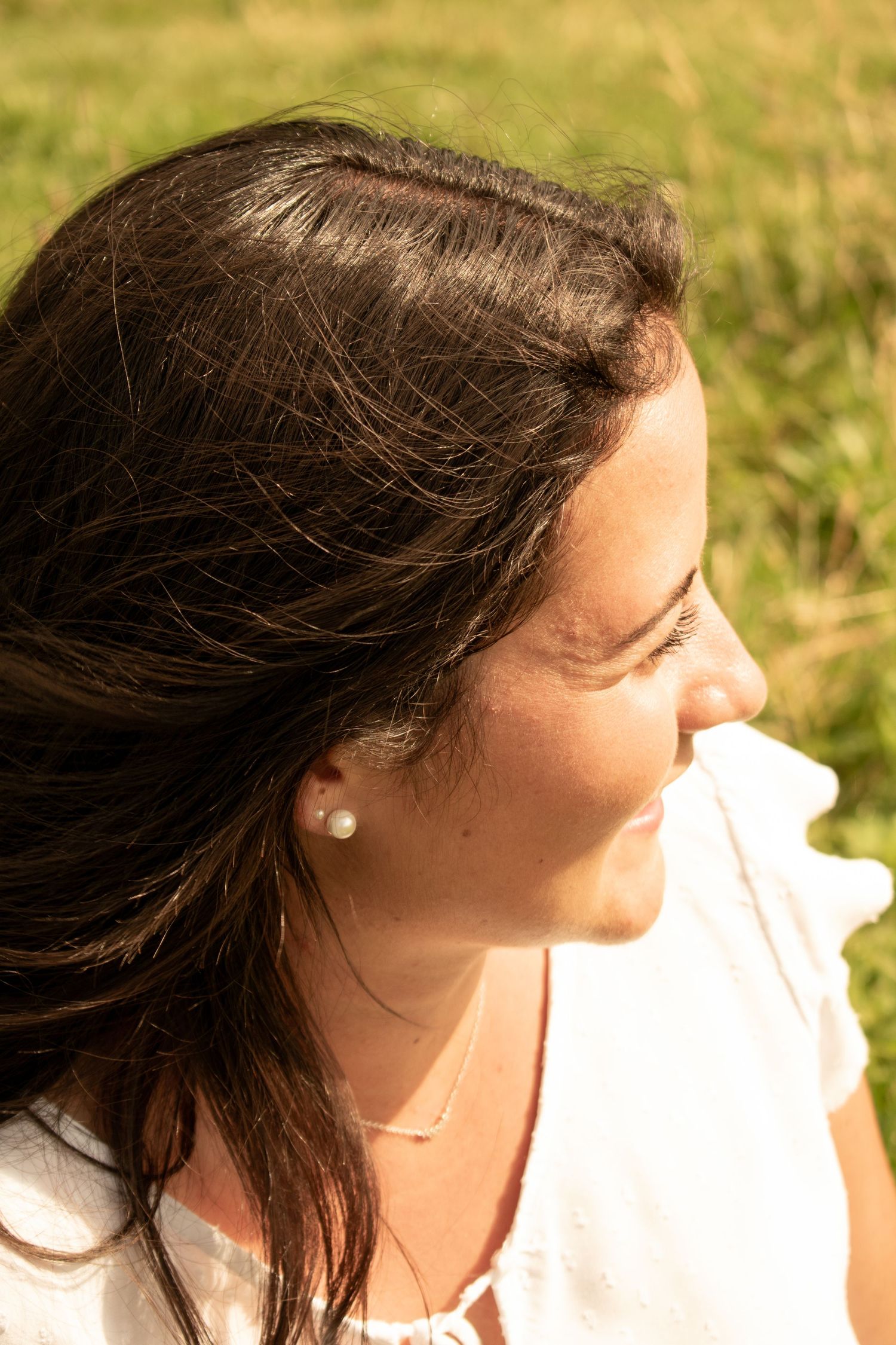 Side profile view of person with pearl earring and necklace smiling in natural outdoor setting.