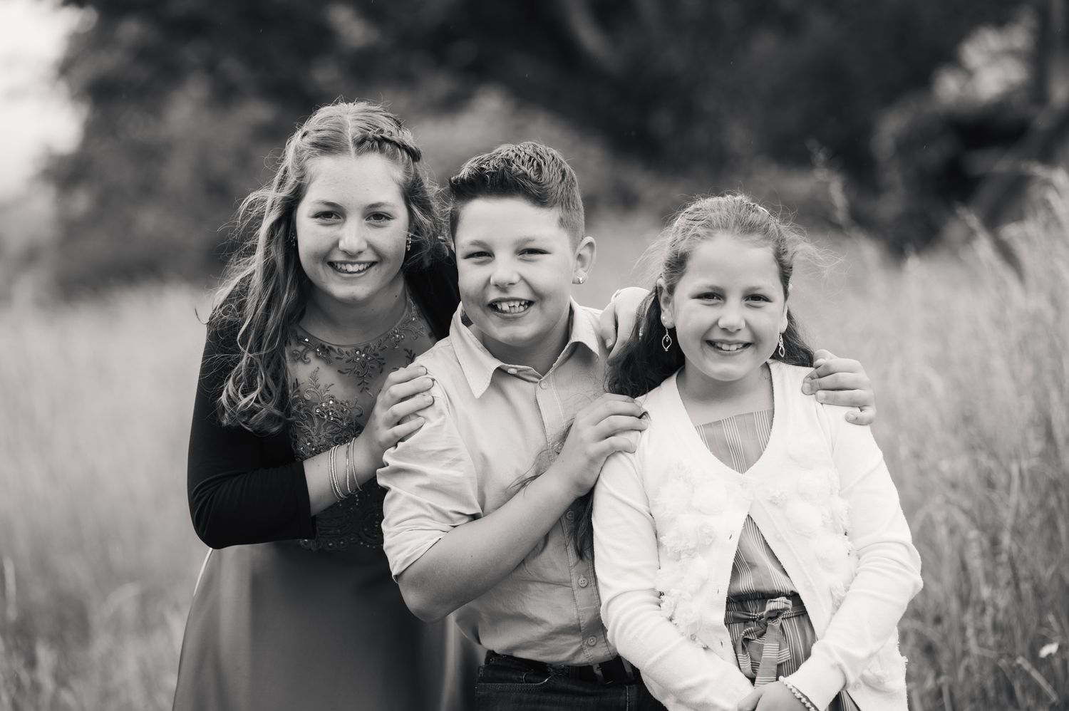 Black and white portrait of three smiling siblings posing together in a field, two girls and a boy in the center.
