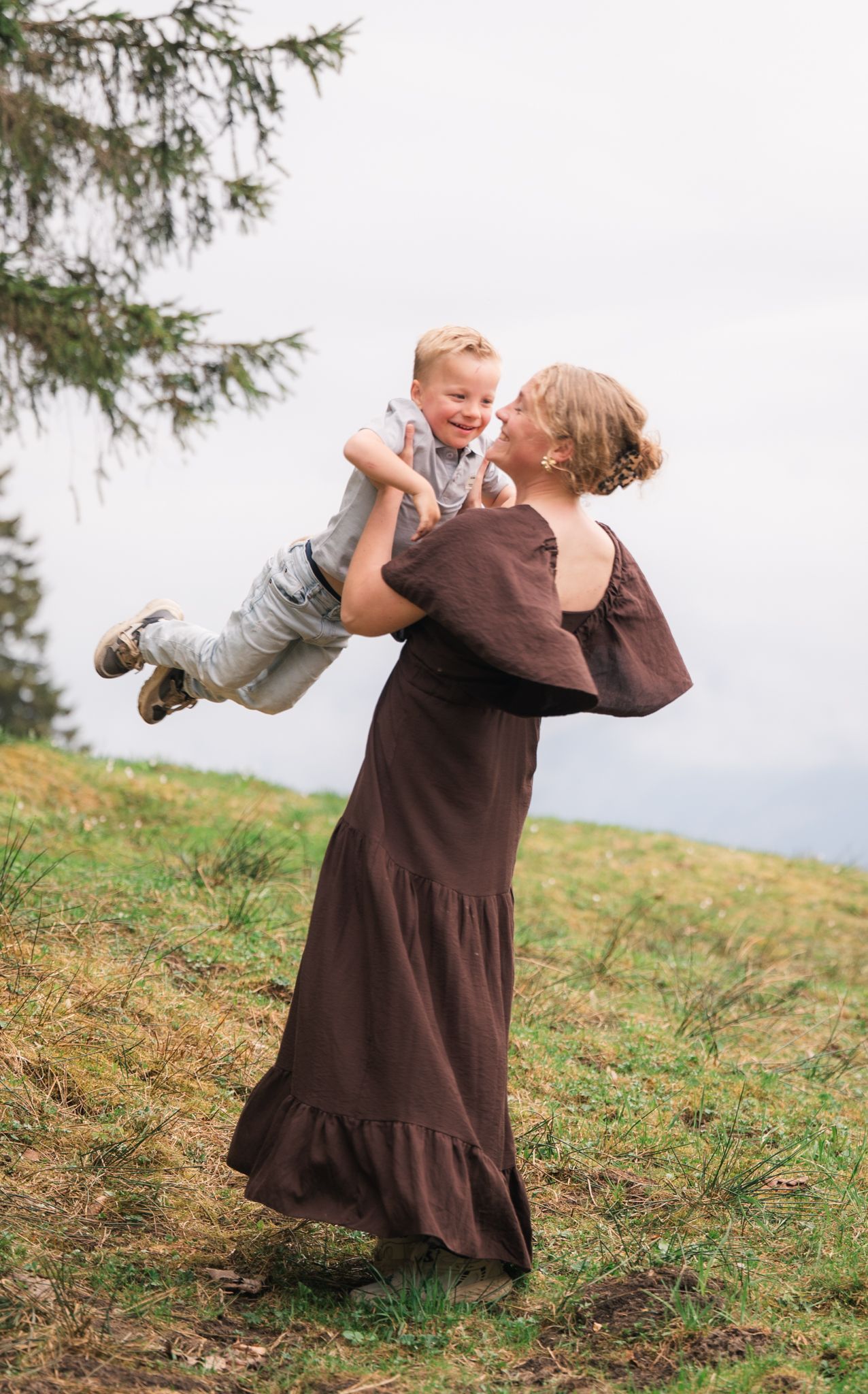 A woman in a brown dress lifts a laughing toddler boy outdoors on a grassy hillside near a pine tree.