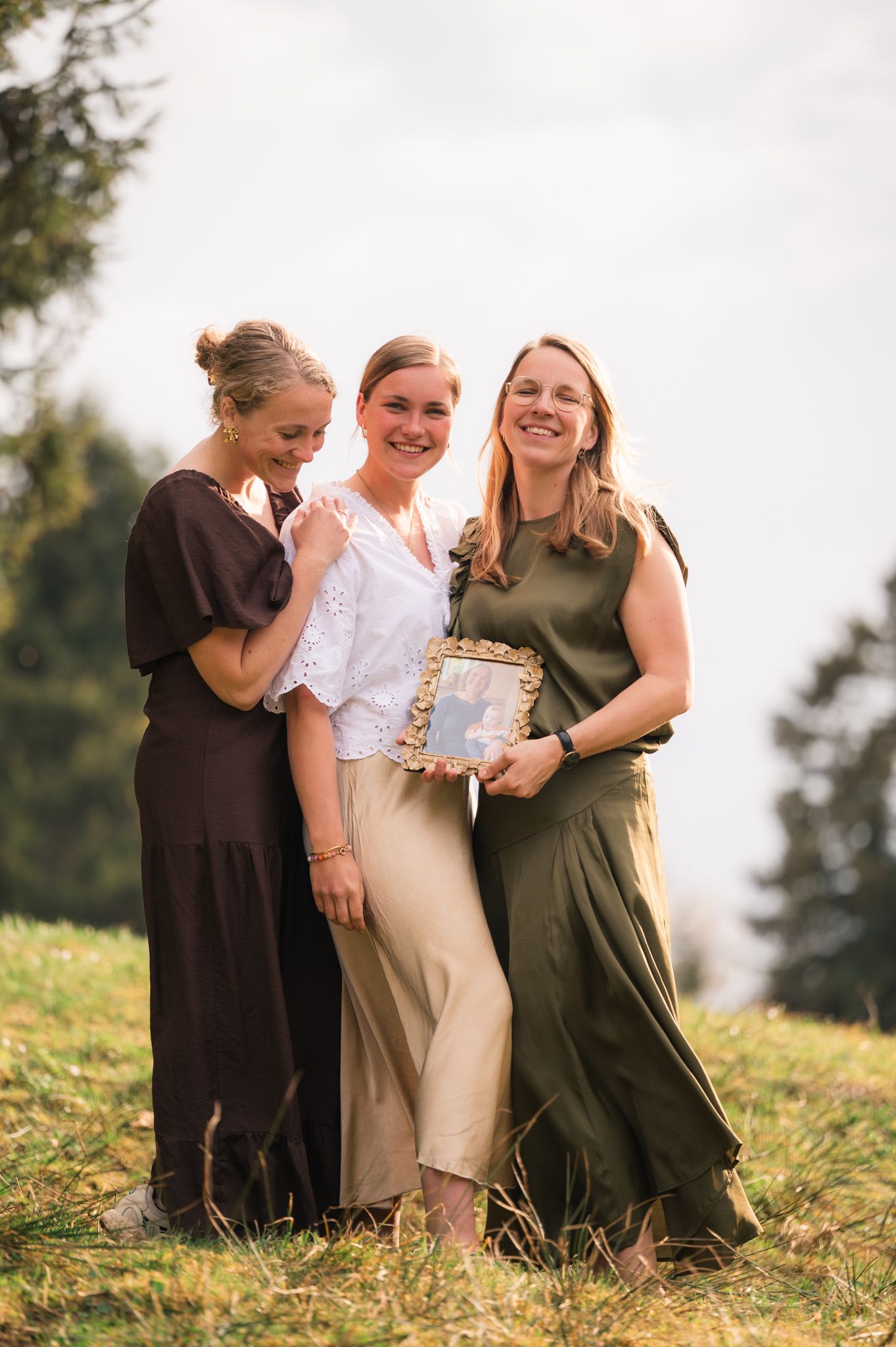Three women smiling outdoors on a grassy hill, one holding a framed photo, dressed in brown, white, and green.