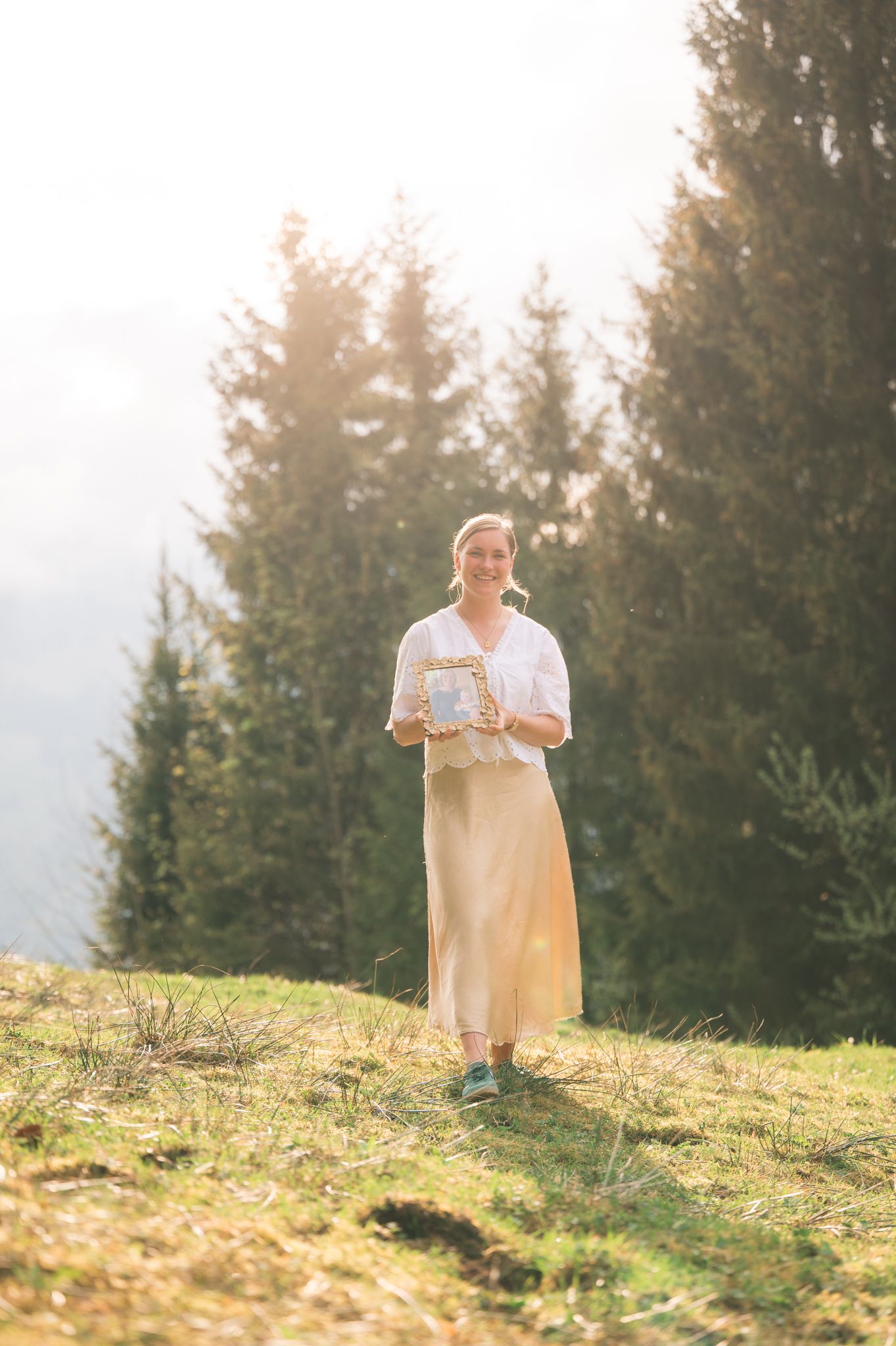 Smiling woman in white blouse and beige skirt holding a gift box on a sunny hillside with evergreen trees.
