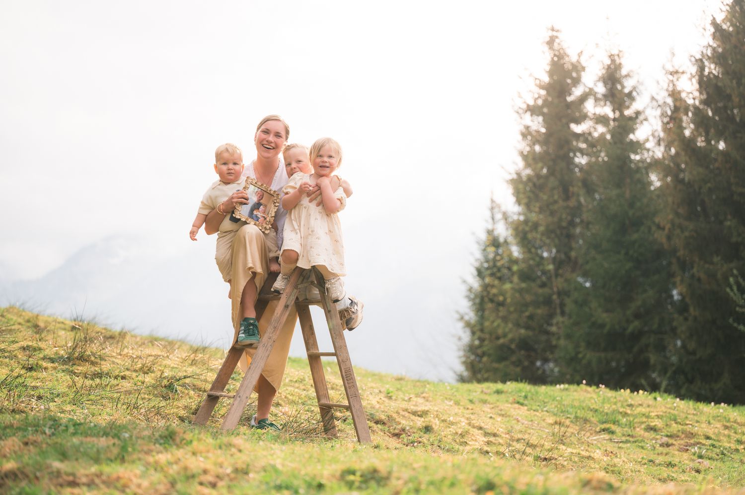 A mother holds two babies on a wooden ladder on a green hillside with evergreen trees and misty mountains behind.