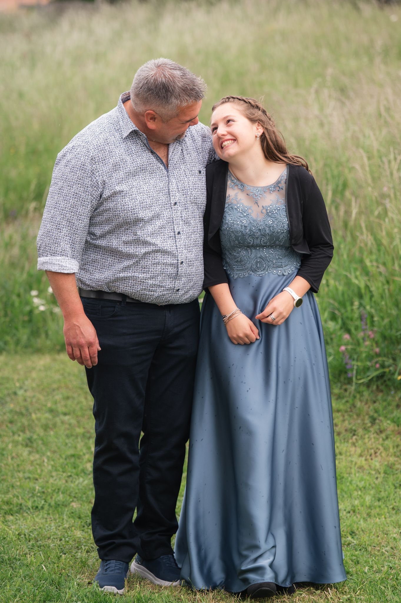 A man and woman share a tender moment outdoors, she wearing a blue gown and black cardigan, standing in a green field.