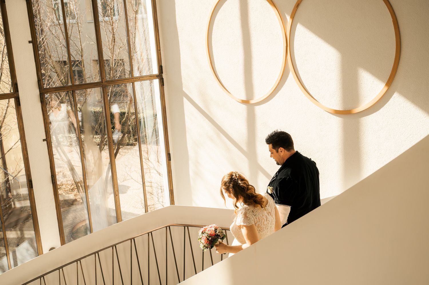 A couple in formal wedding attire stands in a bright sunlit room with large arched windows casting dramatic shadows.