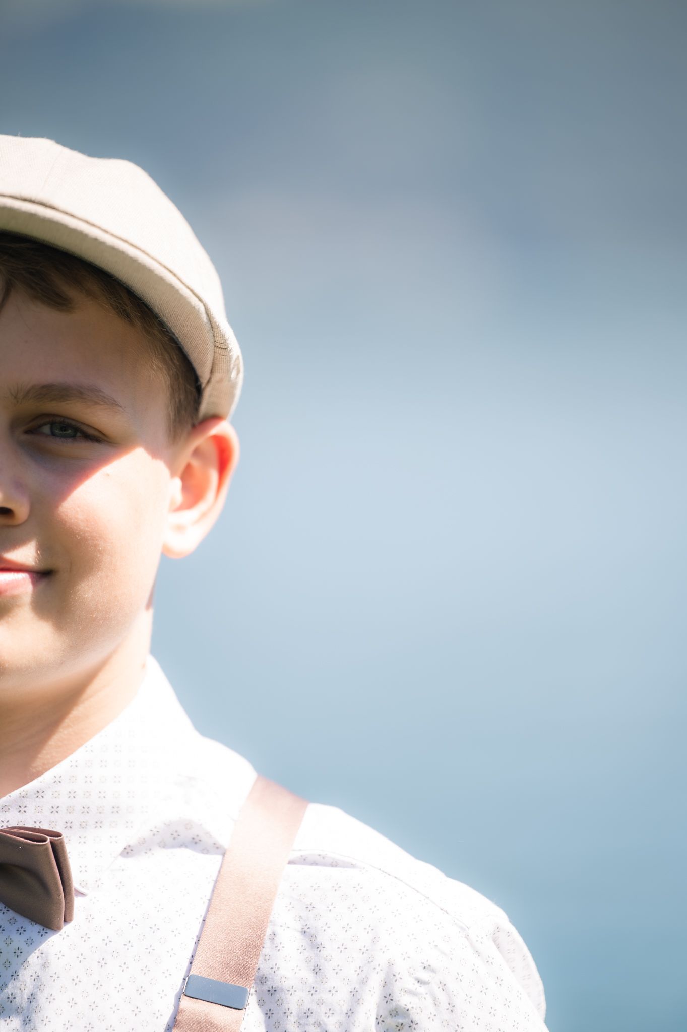 Young boy wearing a flat cap and bow tie smiling outdoors in natural light.