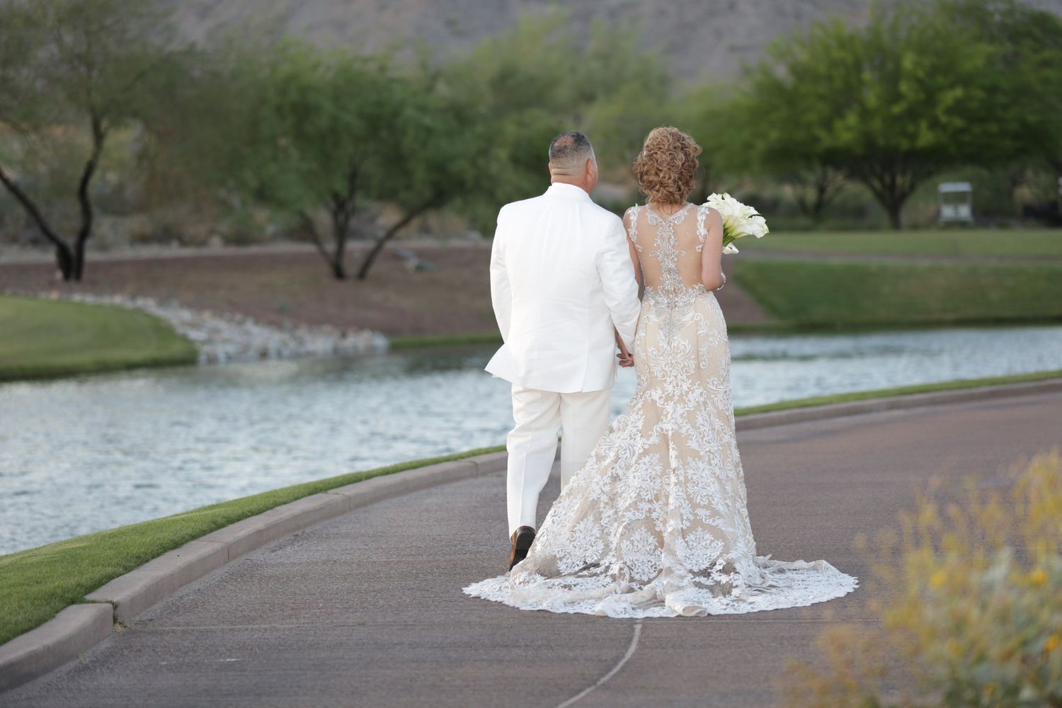 A bride and groom walk together through a scenic outdoor park setting with trees and water in the background.
