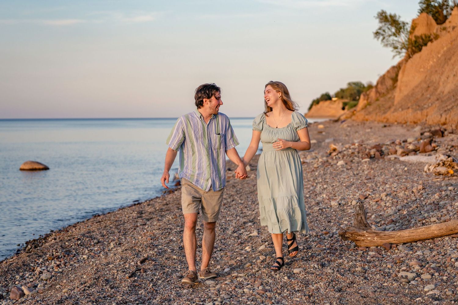 A couple walks along a rocky beach at sunset with golden cliffs and calm ocean waters in the background.