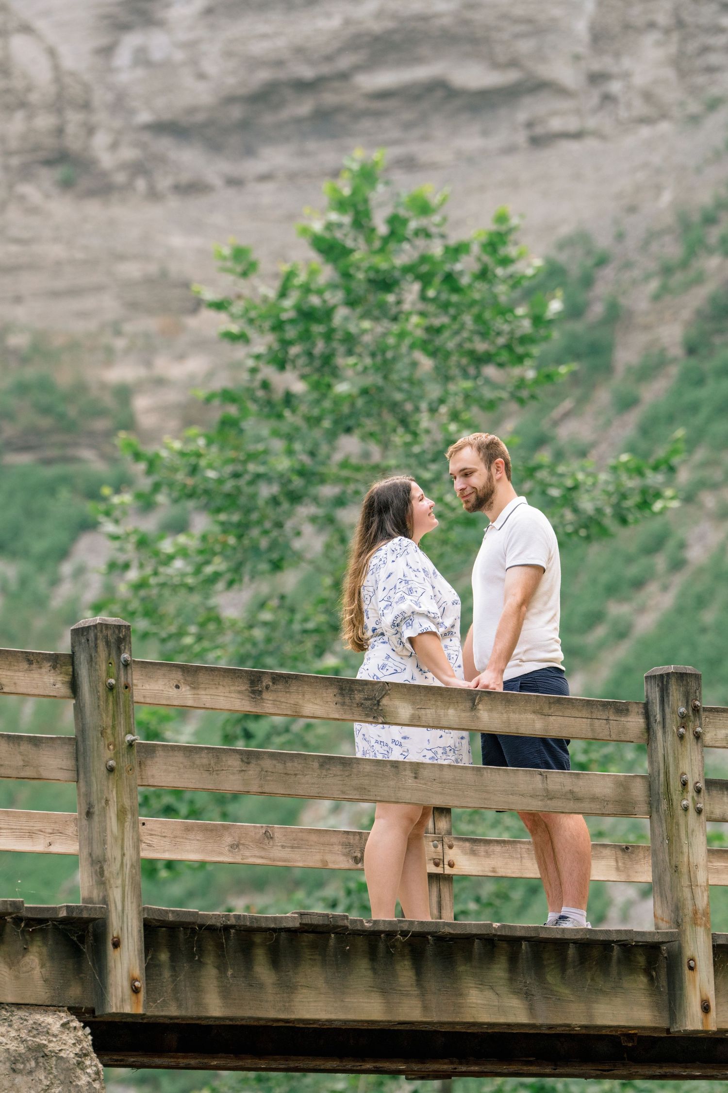 A couple shares an intimate moment while standing on a wooden footbridge with cliffs and greenery behind them.