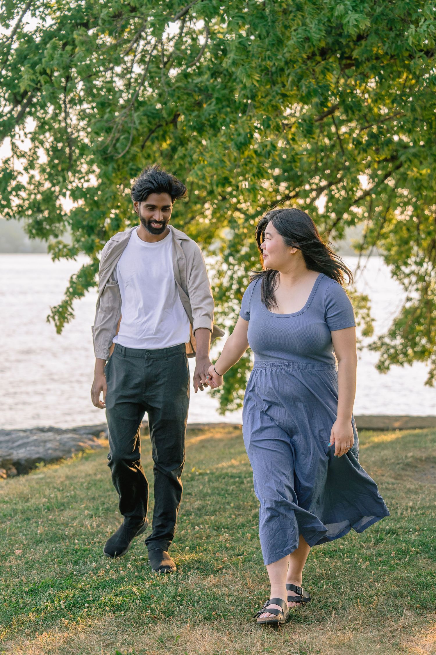A couple playfully dances and spins together on a grassy hill beneath leafy trees.