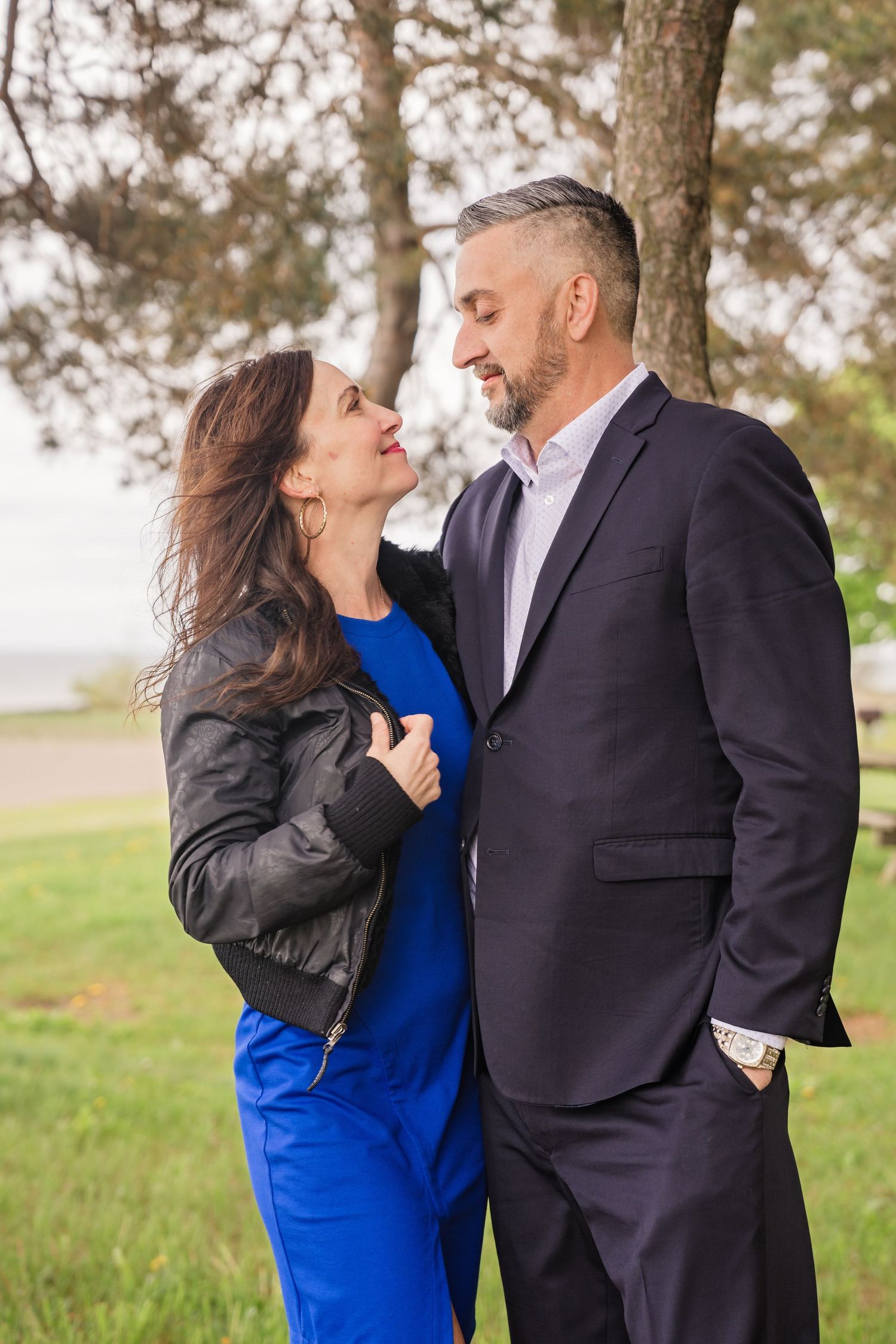 A romantic couple embraces outdoors in a park, with the woman wearing a bright blue dress and man in a dark suit.
