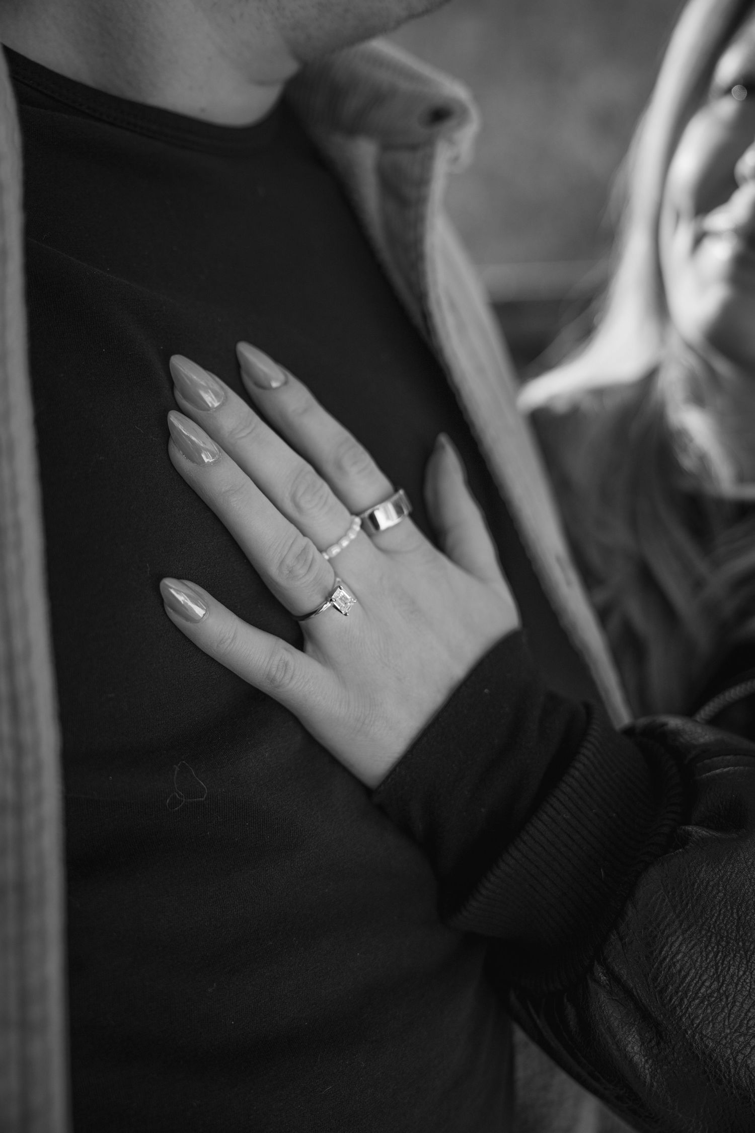 Black and white close-up of a woman's hand with long nails and silver rings resting on a man's chest.