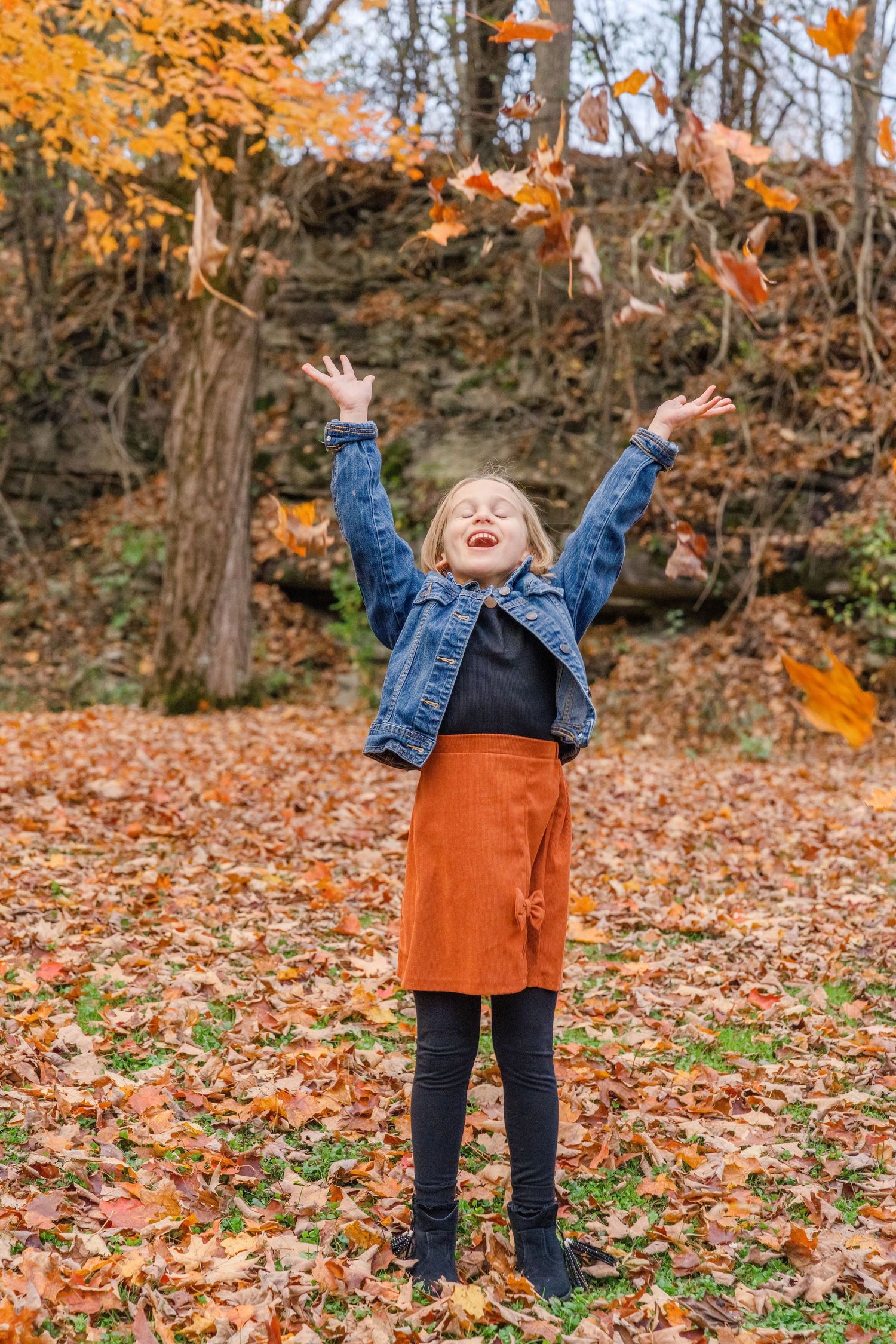 A person in a denim jacket and orange skirt plays joyfully in fallen autumn leaves on a crisp fall day.
