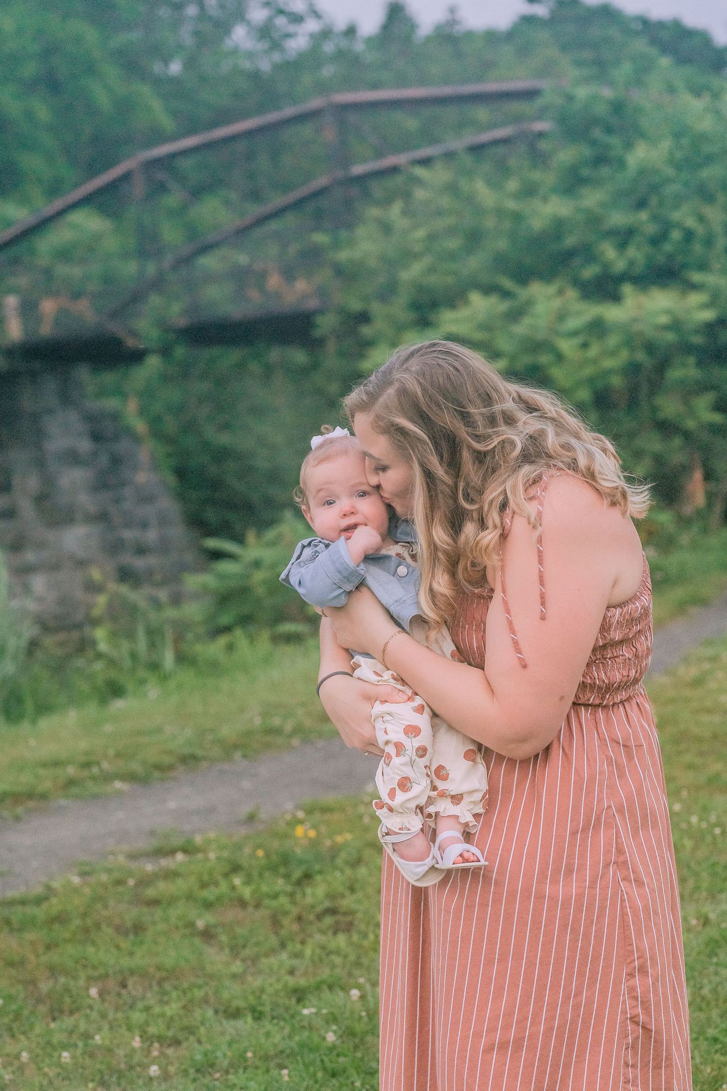 A tender outdoor moment shows a woman in a pink striped dress holding a small baby near a stone bridge.