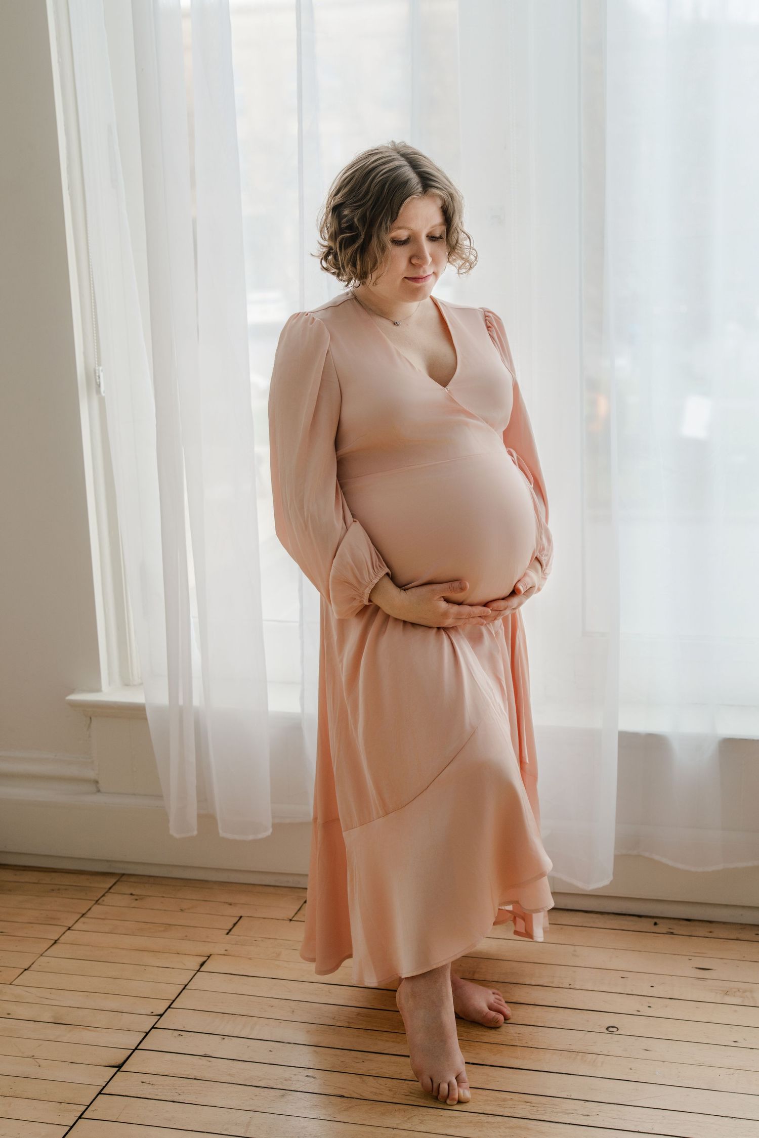 A woman in a flowing peach dress stands by sheer curtains in soft natural light, gently cradling her pregnant belly.