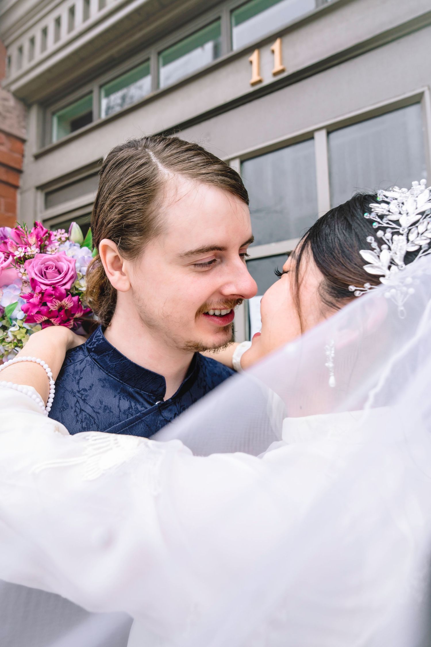 Smiling groom in blue suit embraces bride in white veil and floral headpiece, holding pink bouquet outside building number 11.
